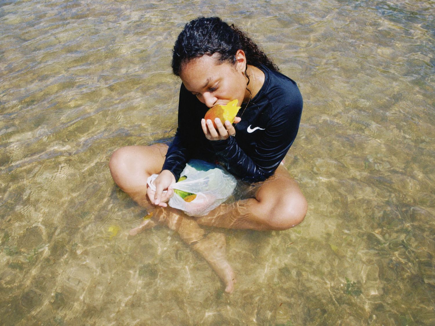 Woman in black Nike shirt sitting in shallow water eating a mango on a tropical beach.