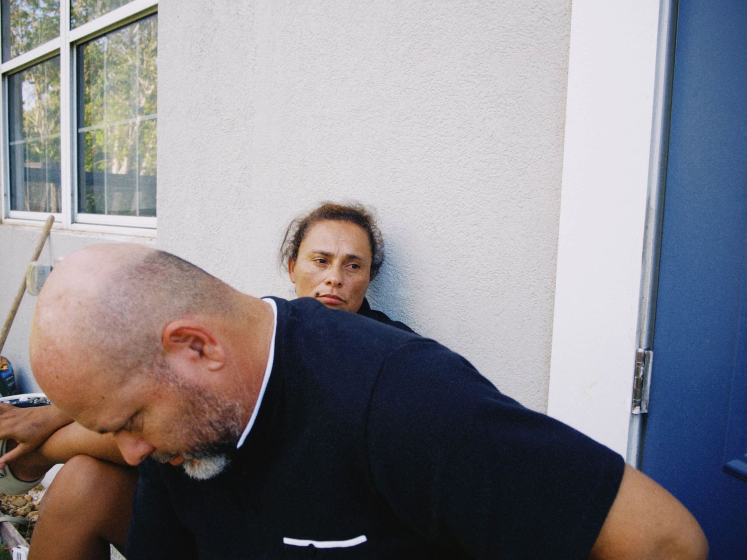 Two men relaxing outdoors against a white building wall, one bald in a black shirt, the other with medium-length hair.