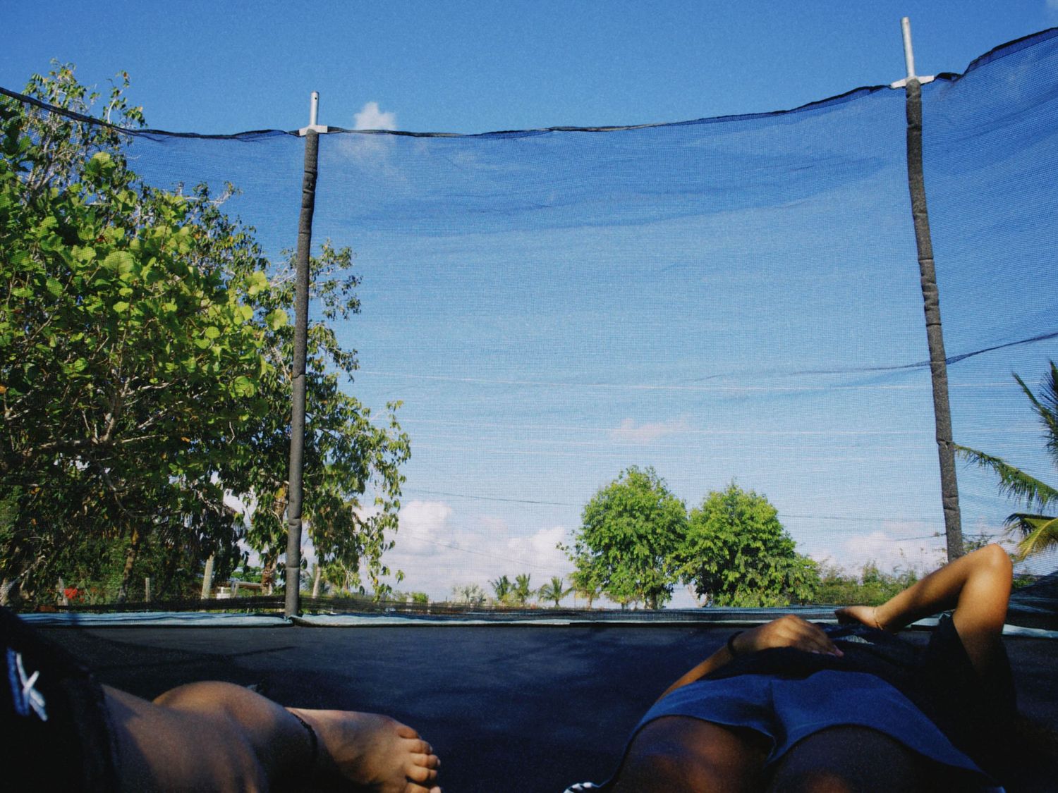 Two people relaxing on a trampoline with safety net, surrounded by green trees under a bright blue sky.