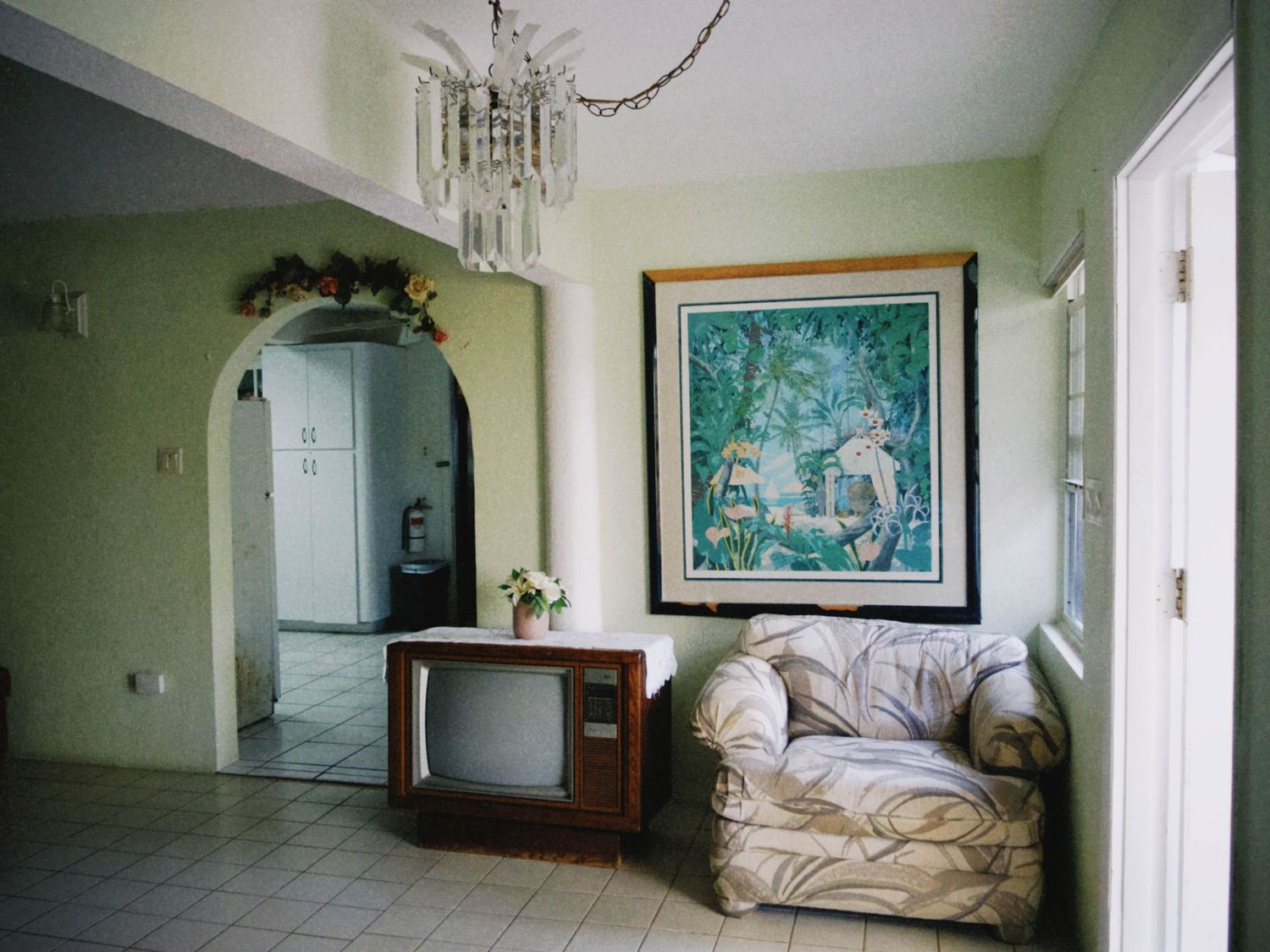 Vintage living room with old TV, floral armchair, large tropical painting, and crystal chandelier over tiled floor.