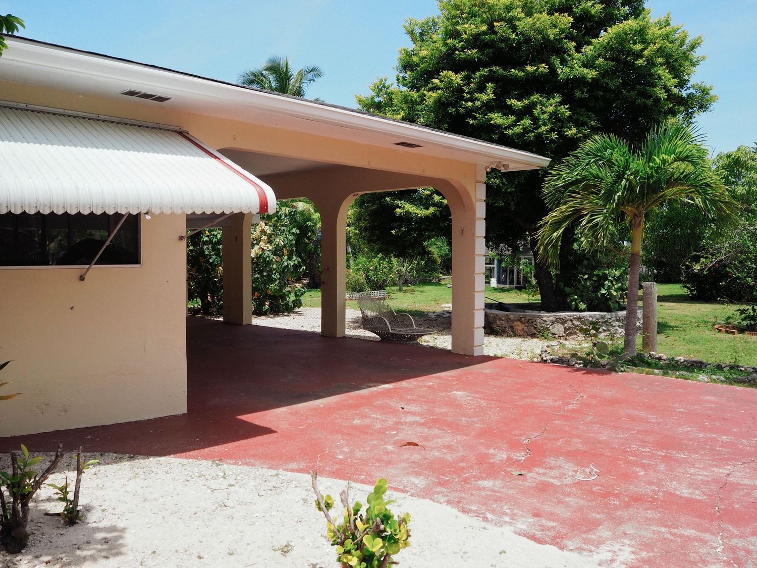 Covered patio with red concrete floor and white columns extends from a house surrounded by tropical palm trees and landscaping.
