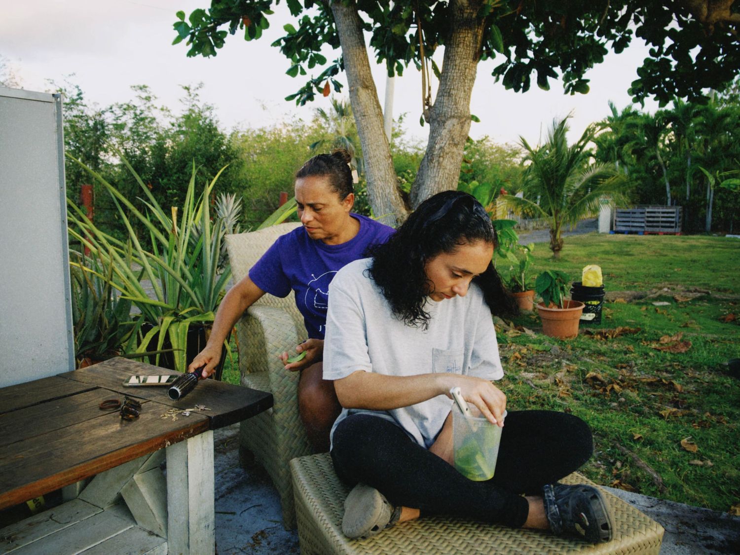 Two women relaxing outdoors near a tree, one reading and one working at a wooden table in a tropical garden.