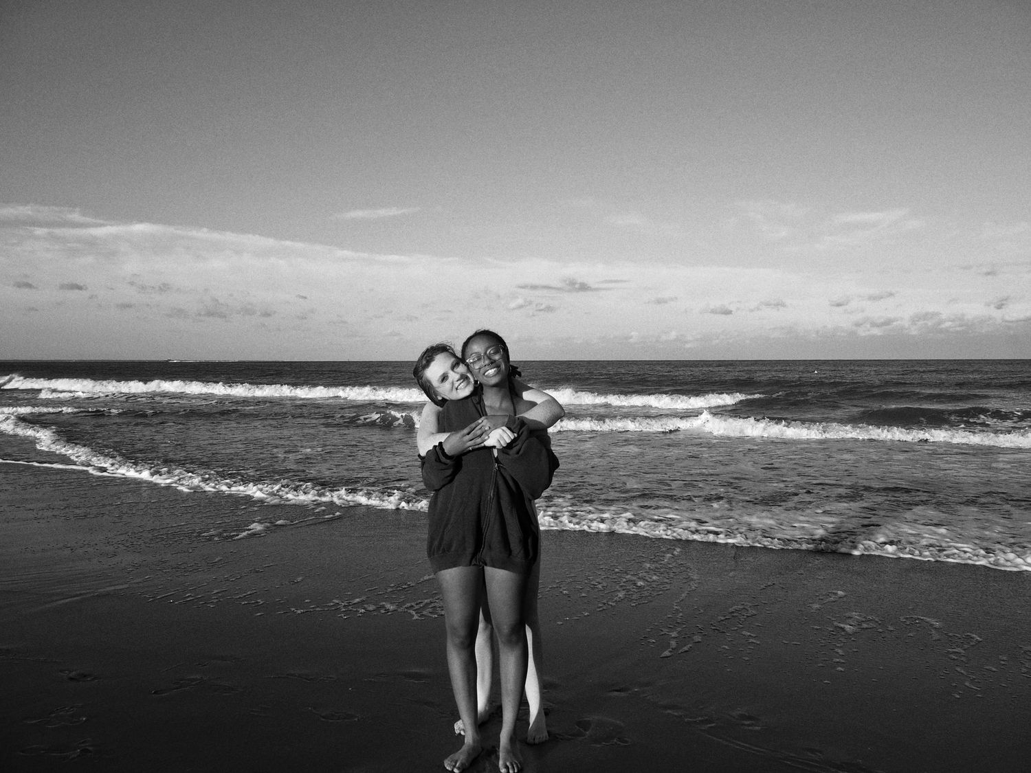 A couple embraces on a black and white beach shoreline with ocean waves and cloudy sky in the background.