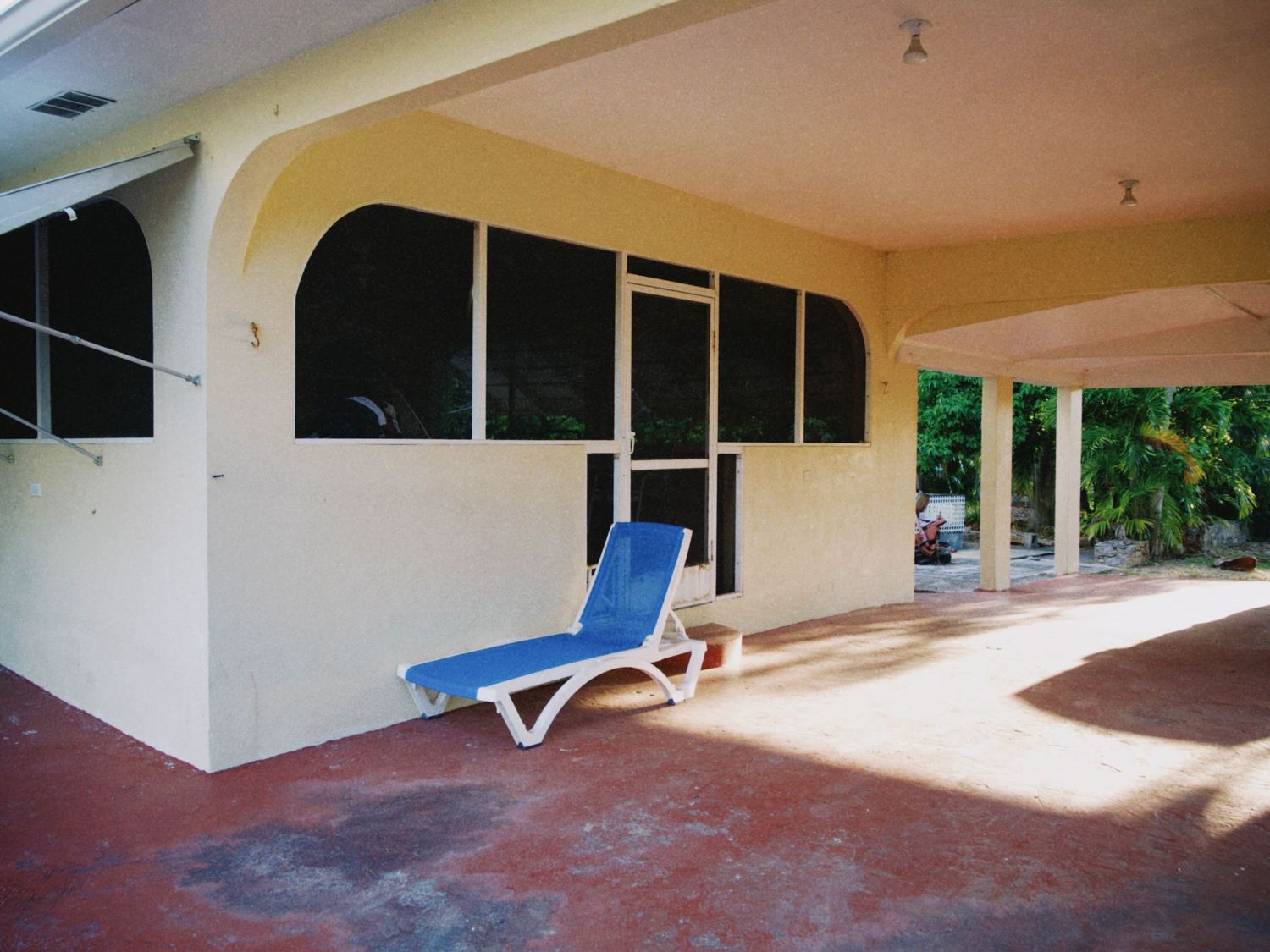 Blue lounge chair on a shaded patio with screened windows and tropical trees in the background.