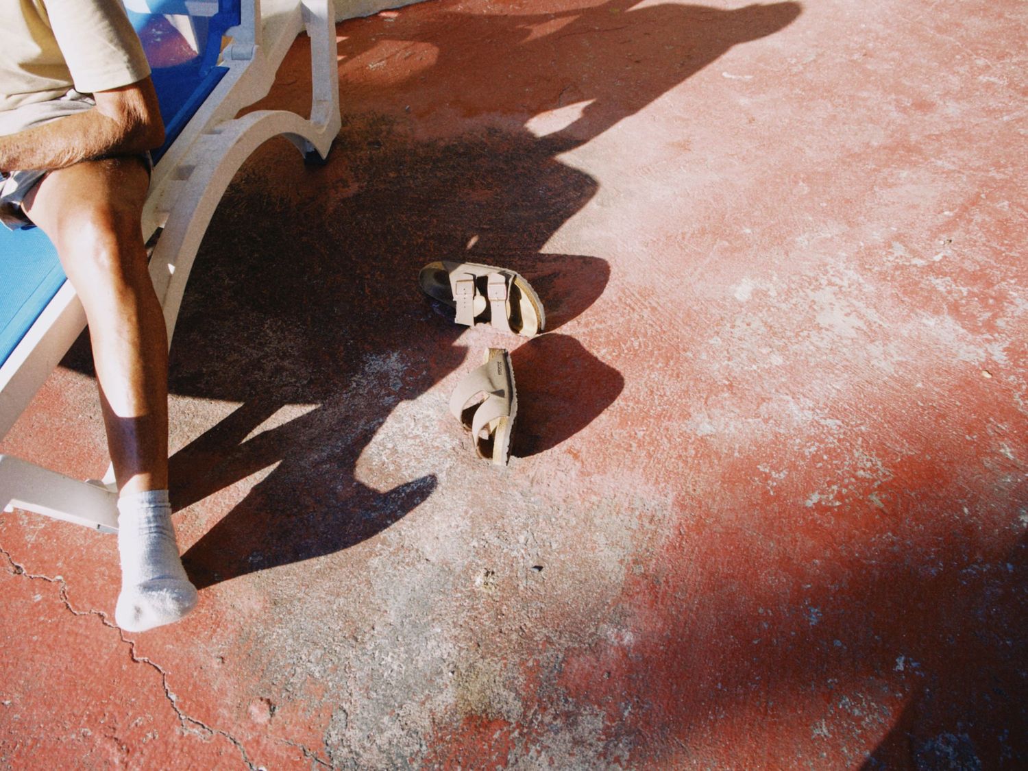 Person sitting on bench with white sandals placed on red ground casting shadows in bright sunlight.