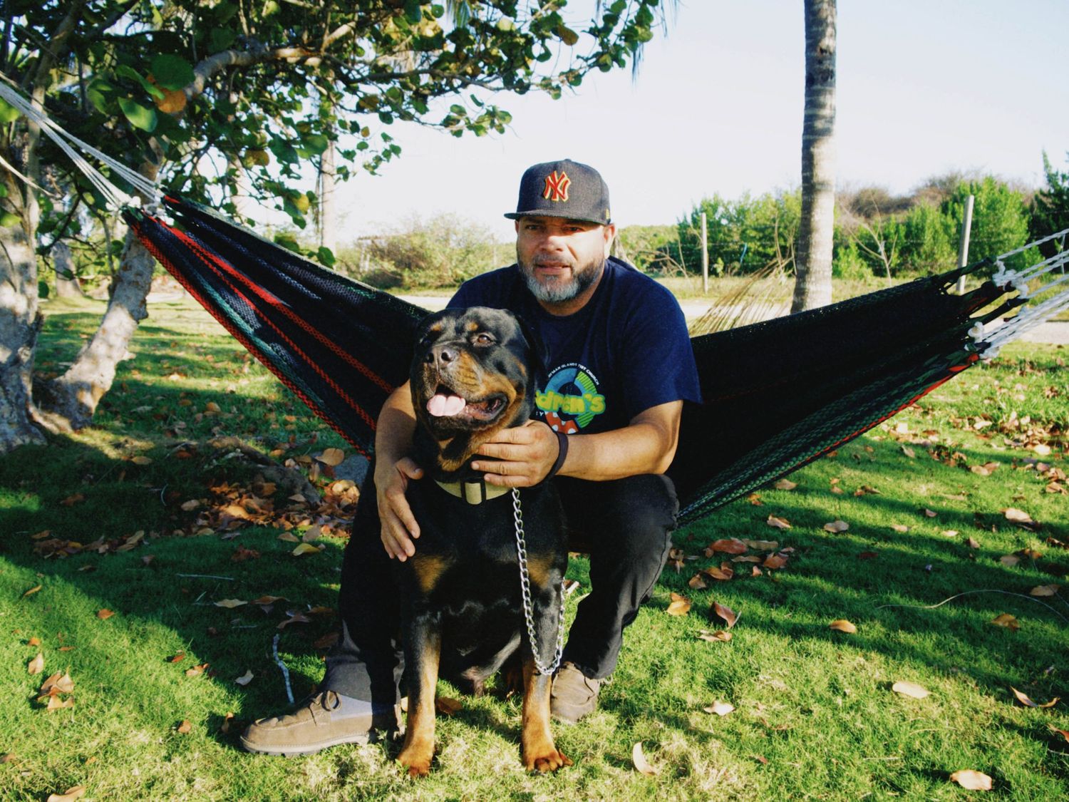 Man in baseball cap hugging his Rottweiler dog while sitting in a black hammock outdoors in a green park.