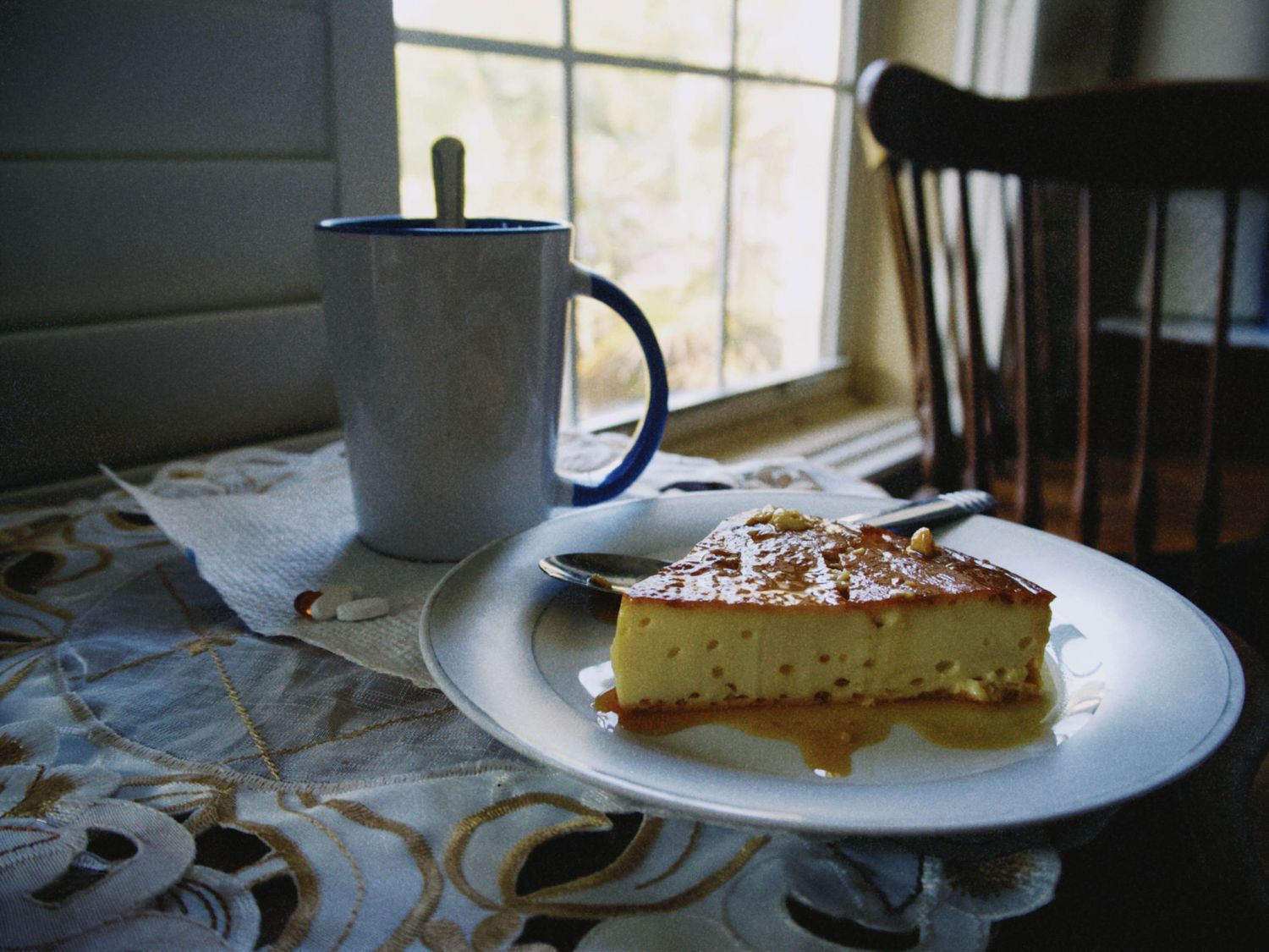 A slice of cheesecake with caramel sauce on a white plate beside a blue mug near a sunlit window.