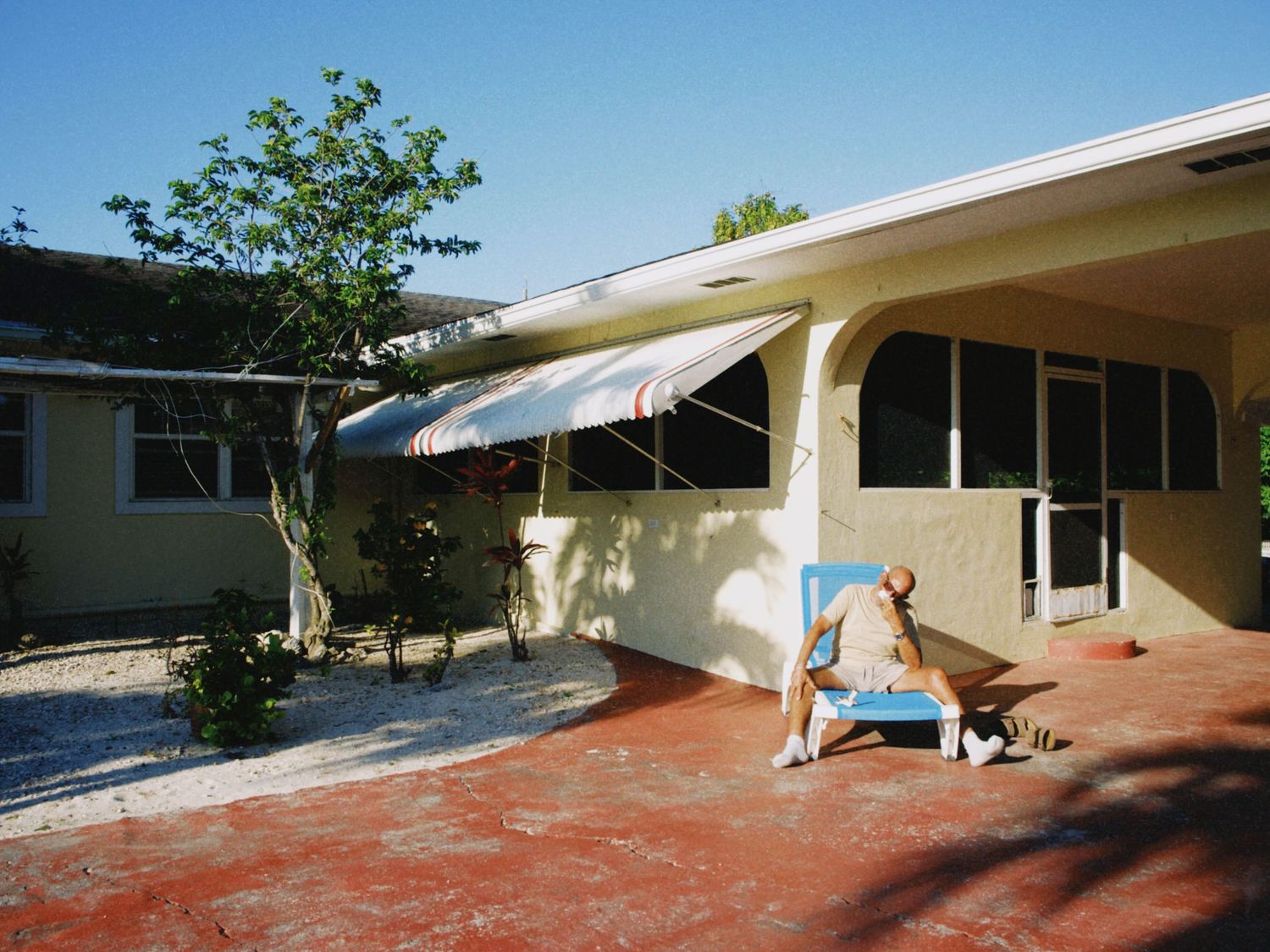 Man kneeling beside a cat outside a Florida-style ranch home with awnings and a brick patio on a sunny day.