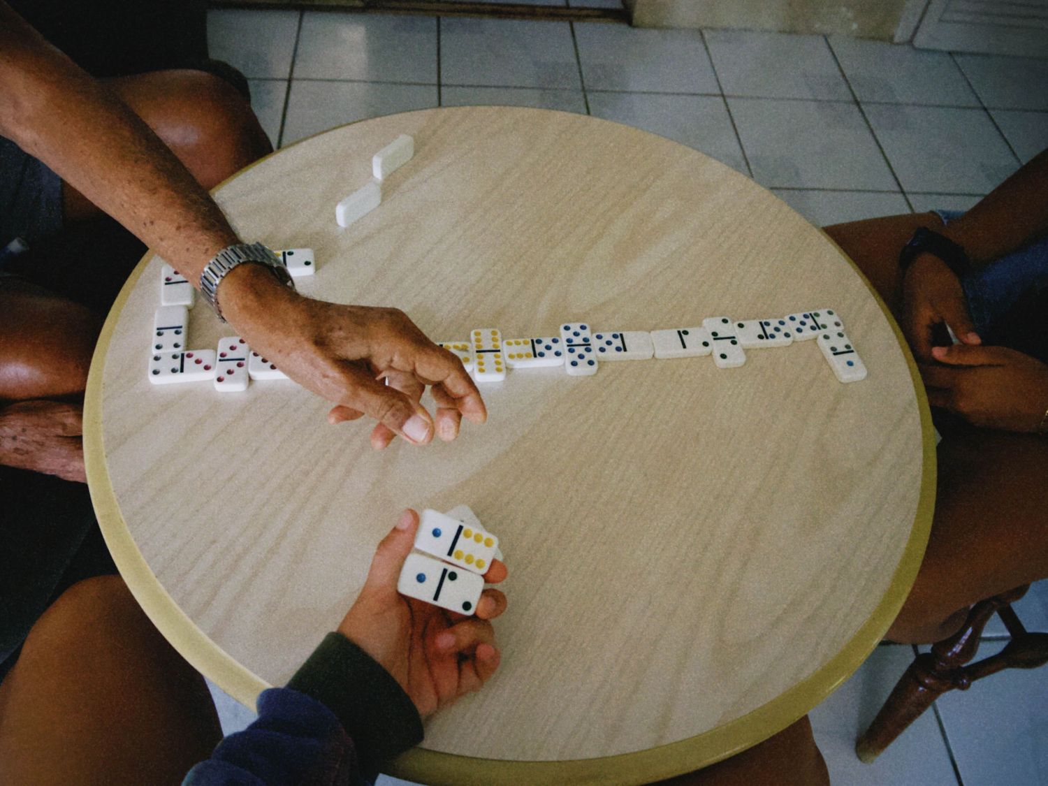 People playing dominoes on a round wooden table, arranging white tiles with black dots in a branching pattern.