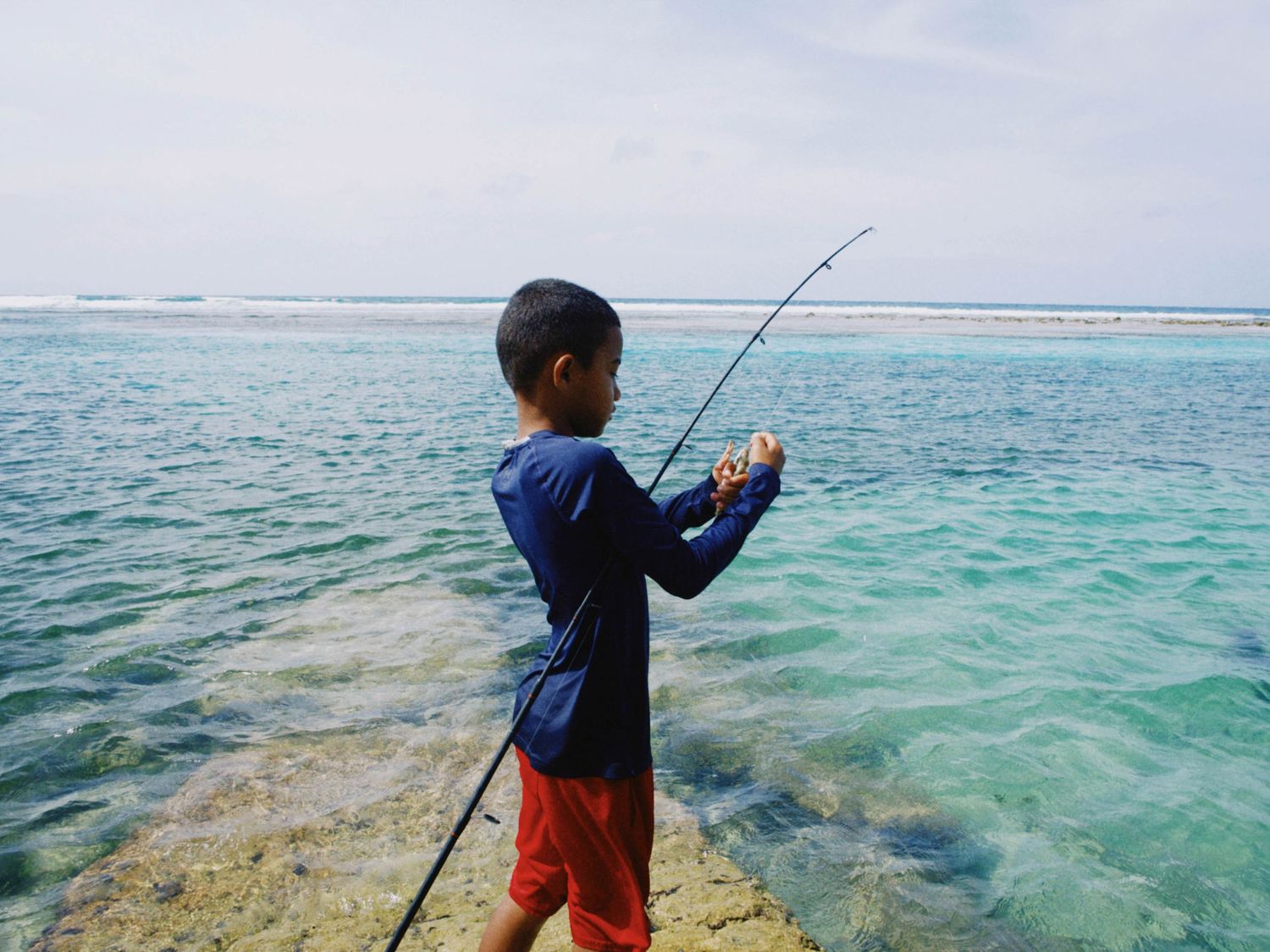 A young boy in a blue shirt and red shorts fishing with a rod by the turquoise ocean shore.