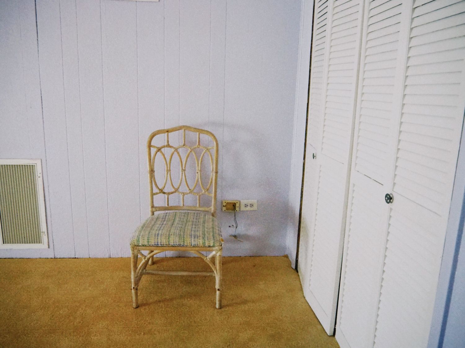Vintage rattan chair with striped cushion in a corner room with white walls and bifold closet doors on beige carpet.