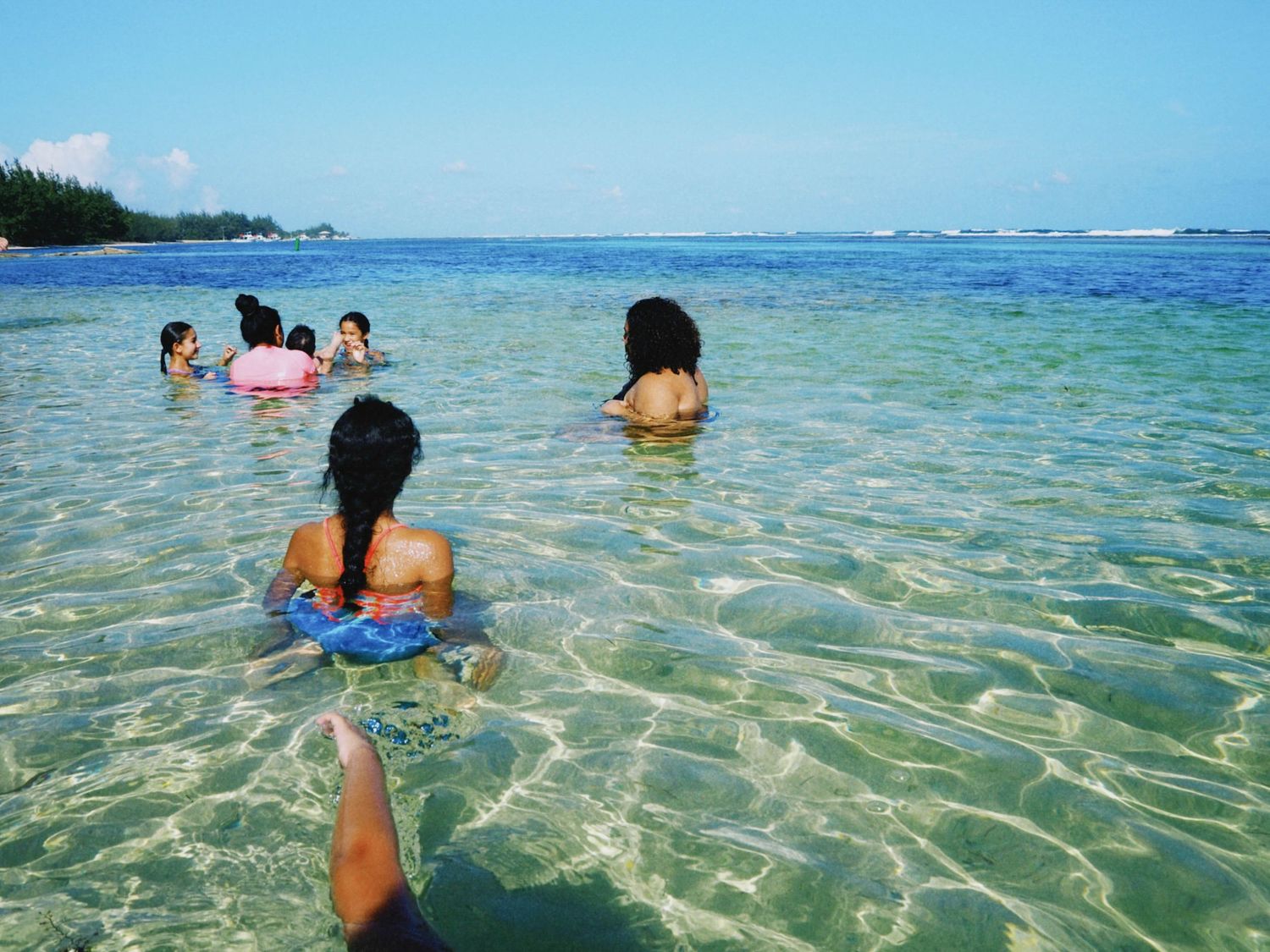 Group of people swimming in clear turquoise tropical ocean waters near a sandy beach with lush green trees.