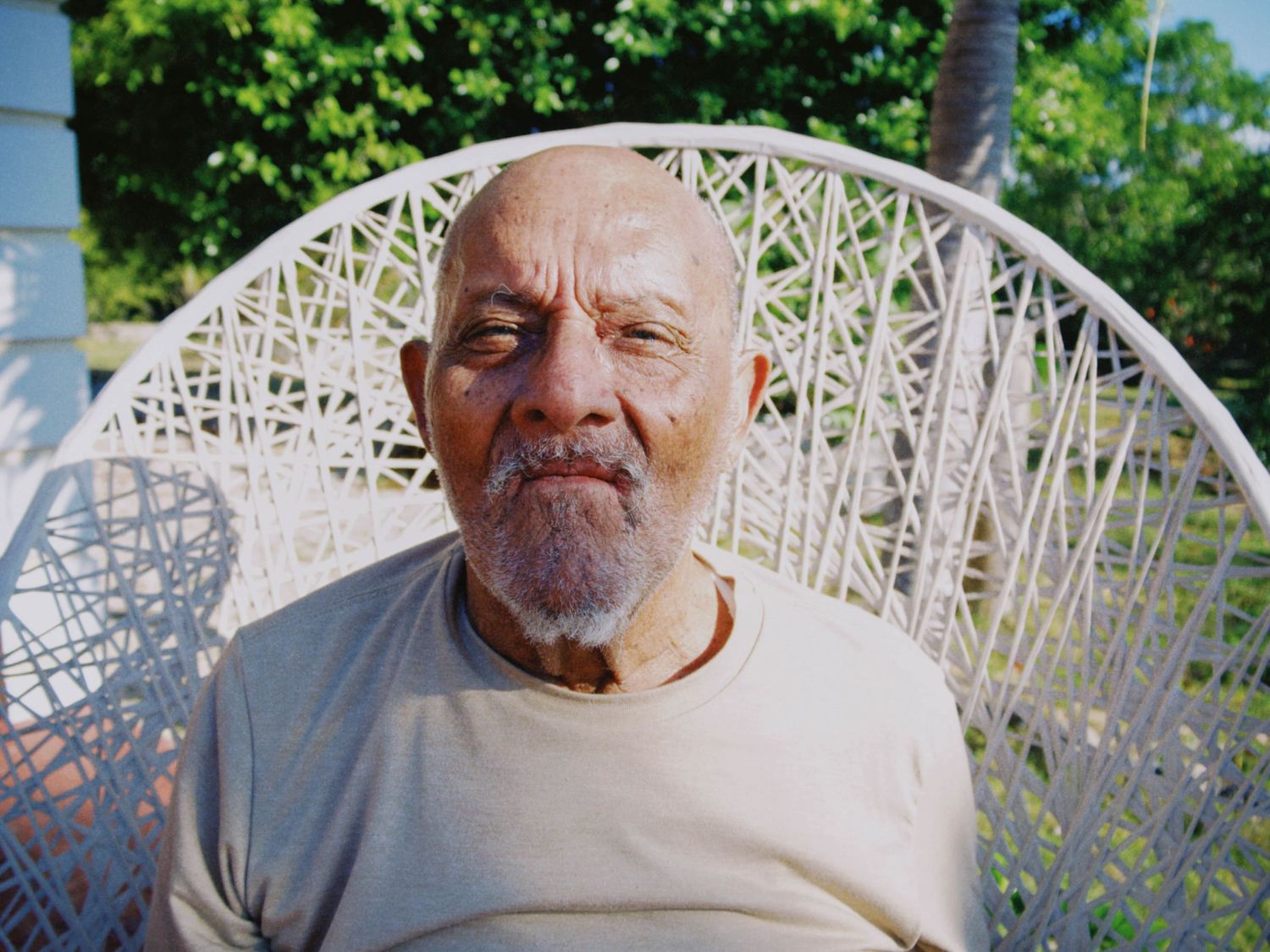 Elderly man with gray beard sitting in white wicker chair outdoors, wearing beige shirt, surrounded by green trees.
