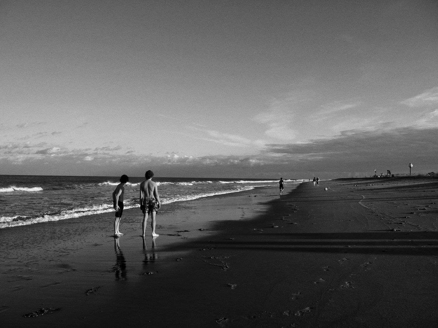 A black and white photograph of two people walking along a beach shoreline with waves and reflections in the sand.