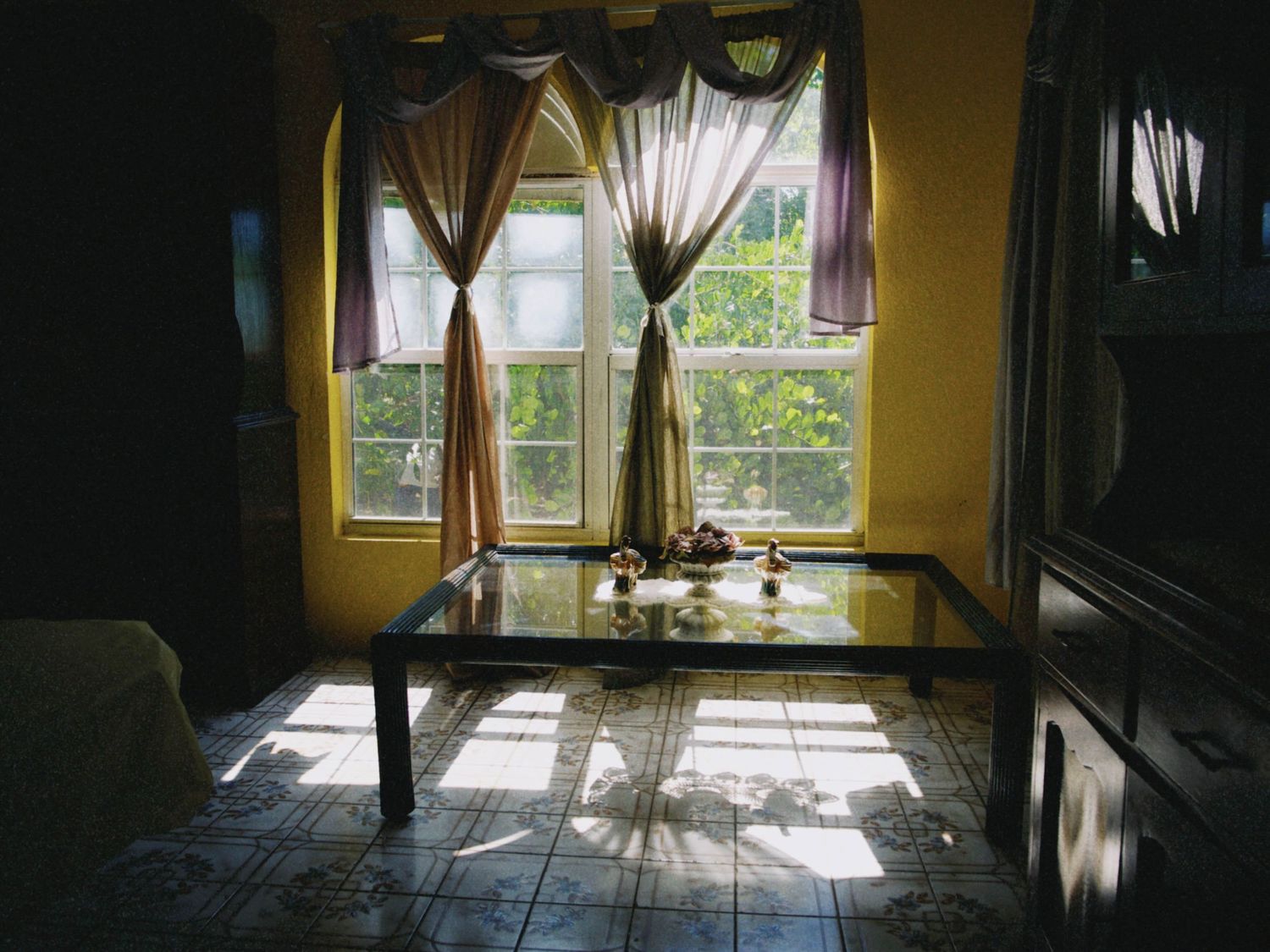 Sunlit dining room with glass table, tiled floor, and elegant draped curtains framing two bright windows.