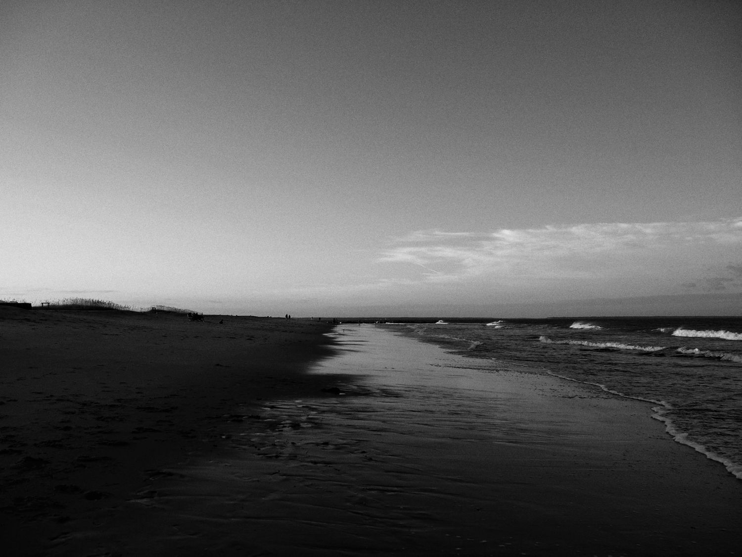 A dramatic black and white seascape with waves rolling onto a dark sandy beach under cloudy skies.