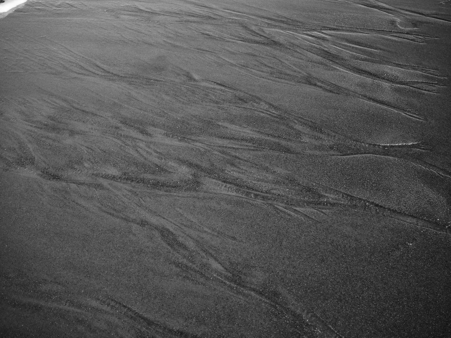 Textured ripples and patterns in dark wet sand created by water flow on a beach surface.