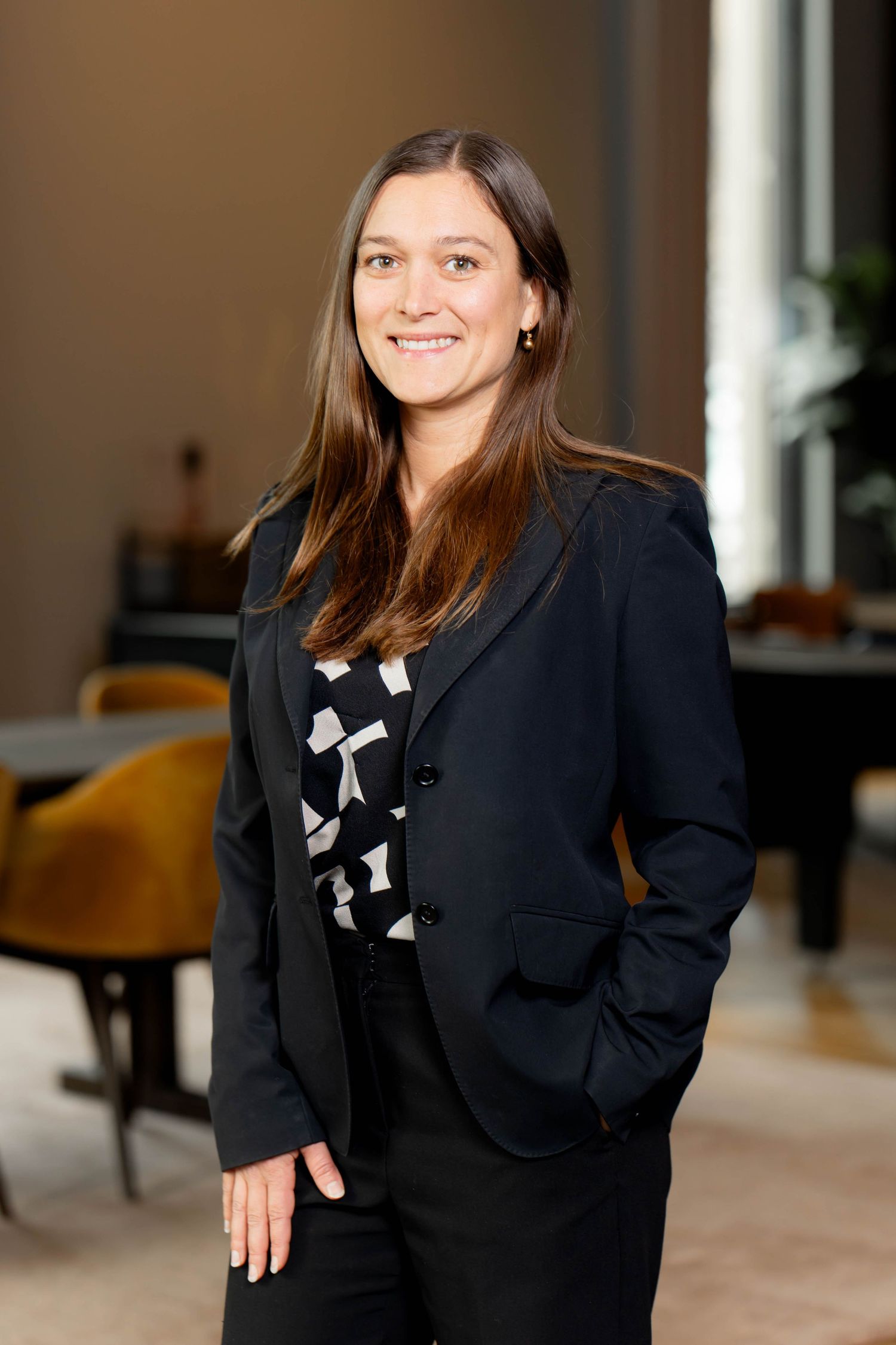 Professional portrait of someone in a black blazer and patterned top smiling warmly in an office setting.