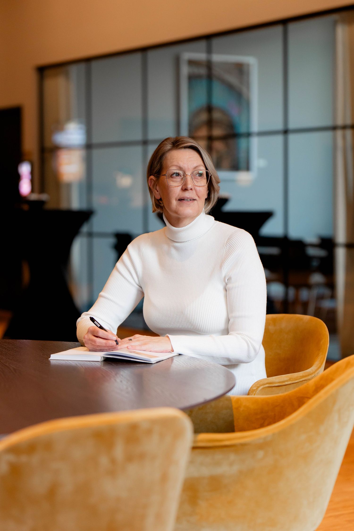 Person in white turtleneck sweater sits at wooden table in modern office space with blurred background.
