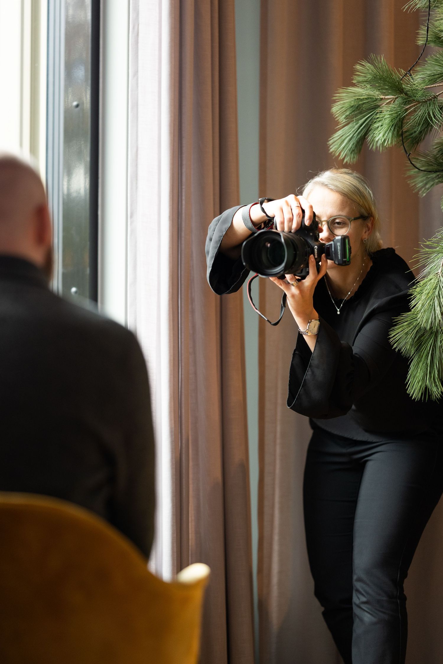 Professional photographer in all black taking photos near a window with natural light and decorative pine branches.