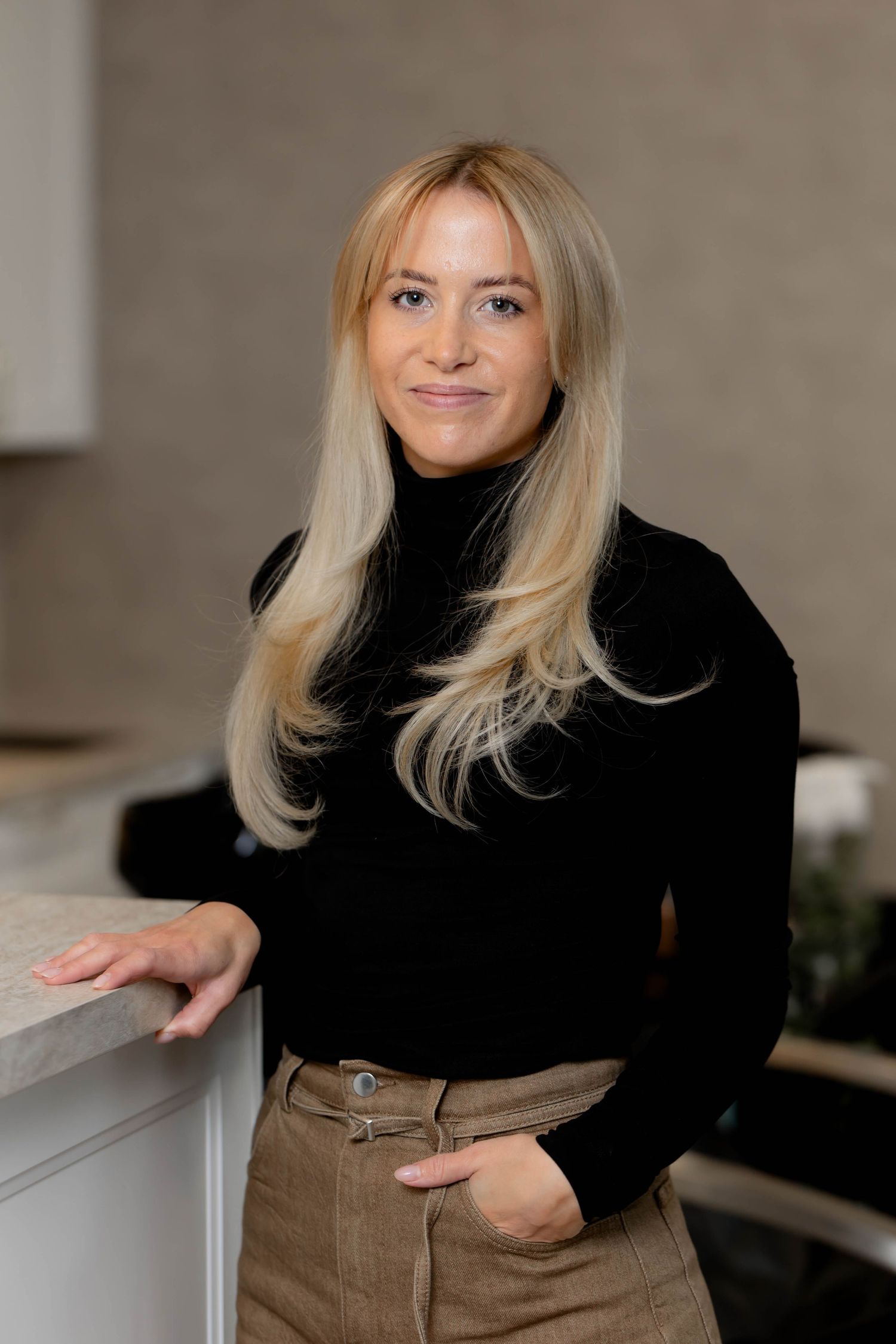 Professional portrait series of person in black turtleneck and brown pants leaning against counter in modern interior.