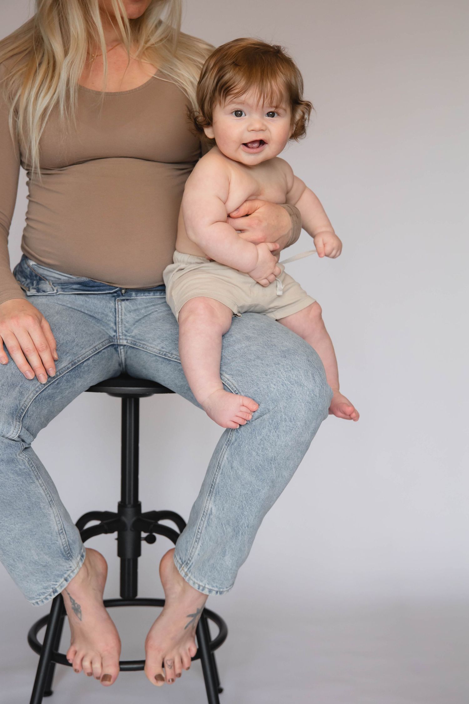 Smiling baby sitting on mother's lap on a black stool, wearing beige shorts, with mom in jeans and tan top.