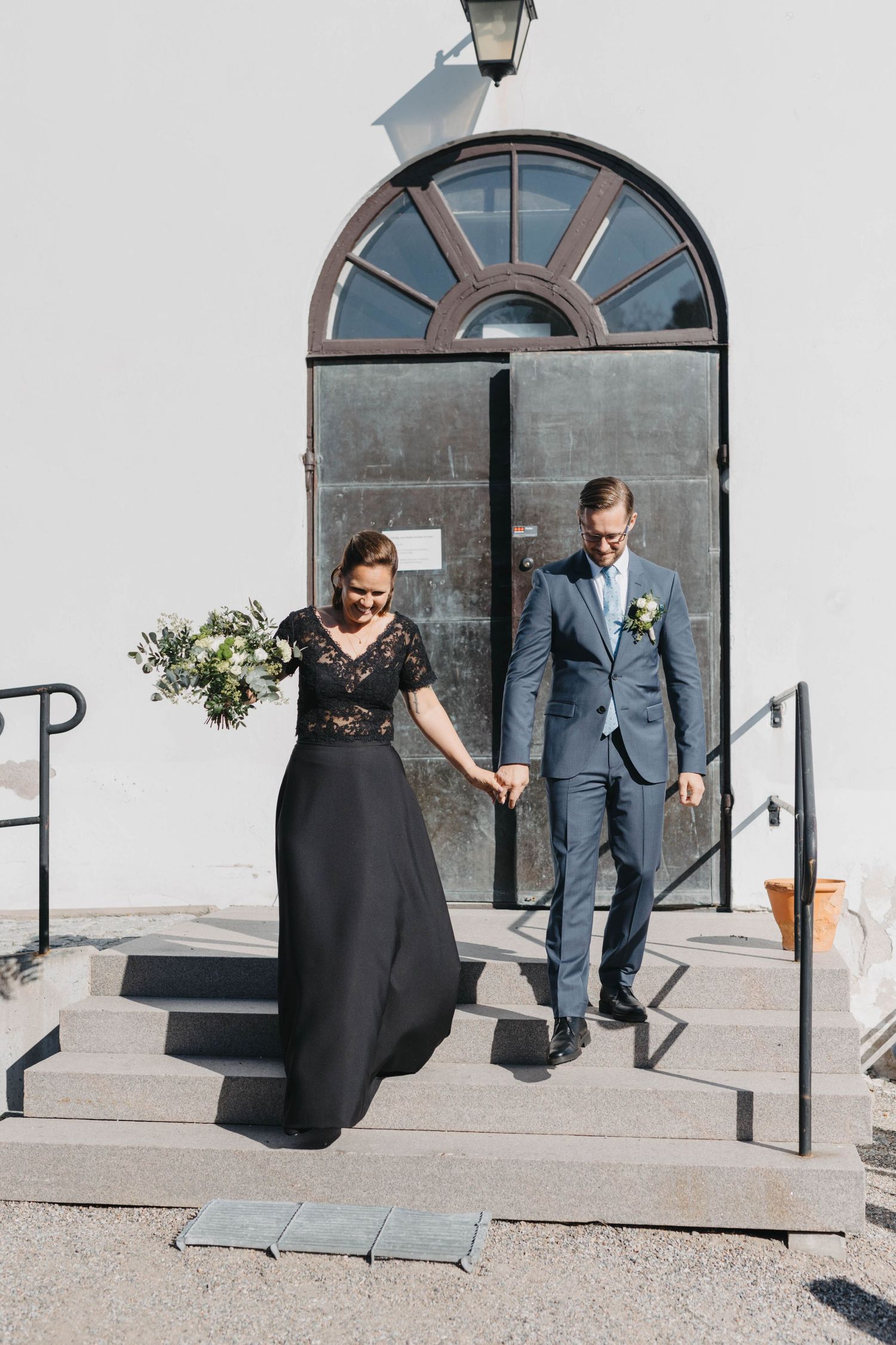 A couple in formal attire descends stone steps outside a building with an arched doorway and vintage lamp.