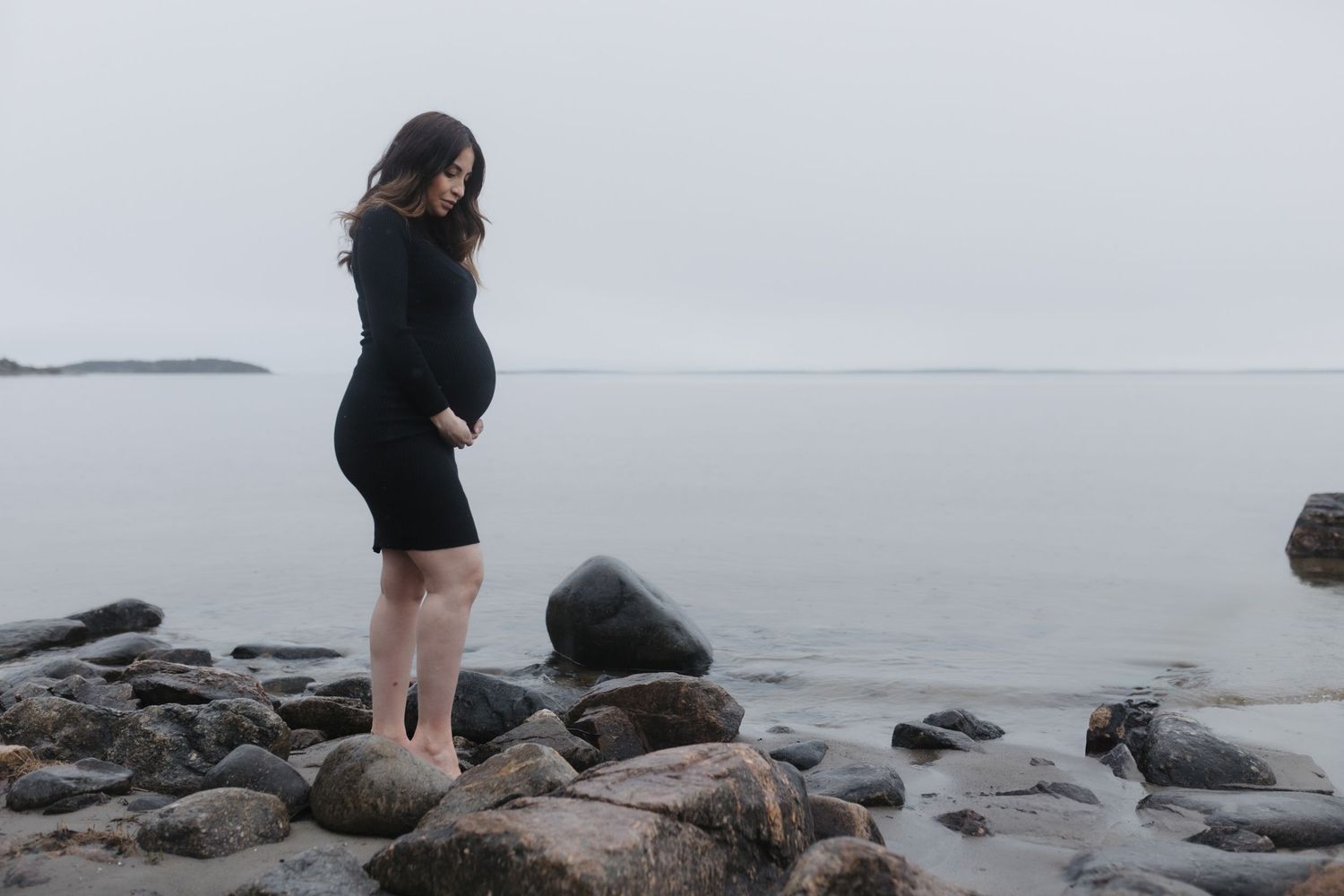 A person in black clothing stands on rocky coastal terrain overlooking a misty gray ocean.