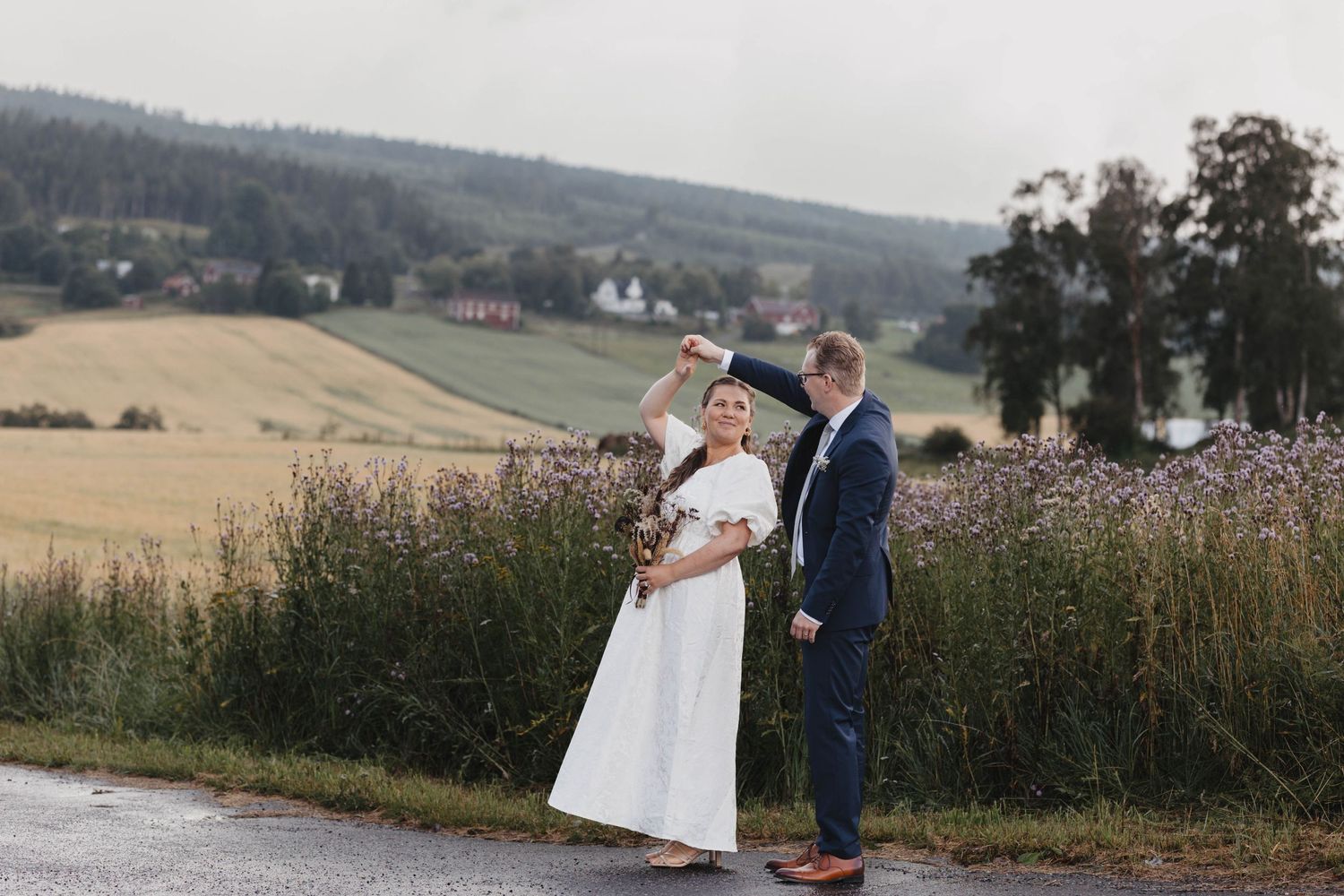 Newlywed couple embraces in a scenic rural field with rolling hills and farmland in the background.