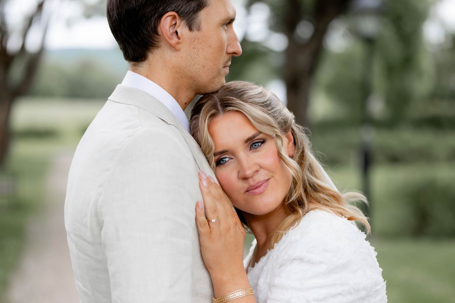 A tender moment between a couple in white attire embracing outdoors with a soft natural backdrop.