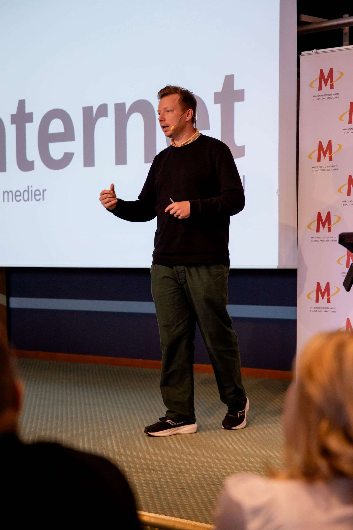 A presenter in black clothing gestures while giving a talk at a business conference with Medipr branding on display.