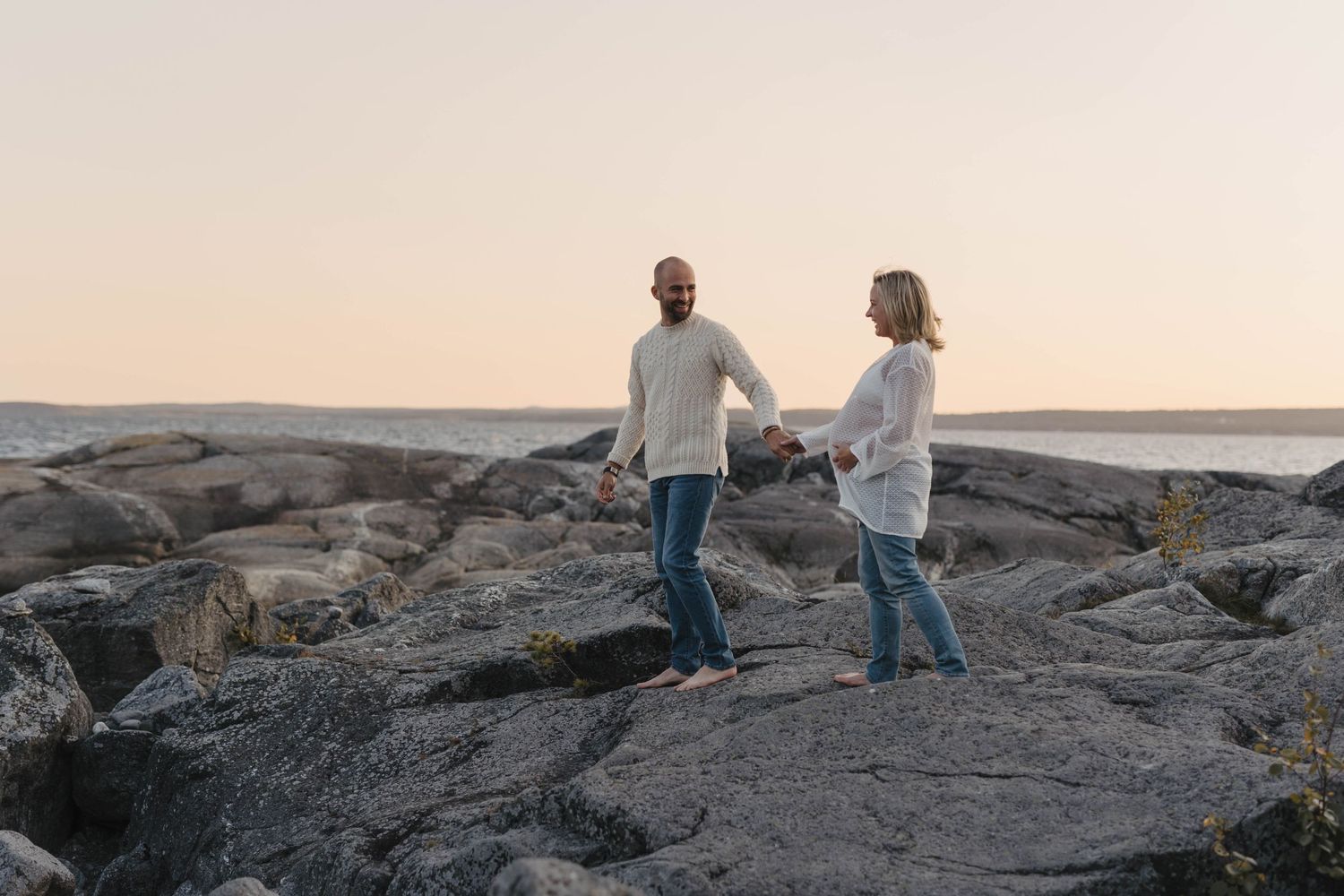 A couple holds hands while walking on rocky coastal terrain at sunset.
