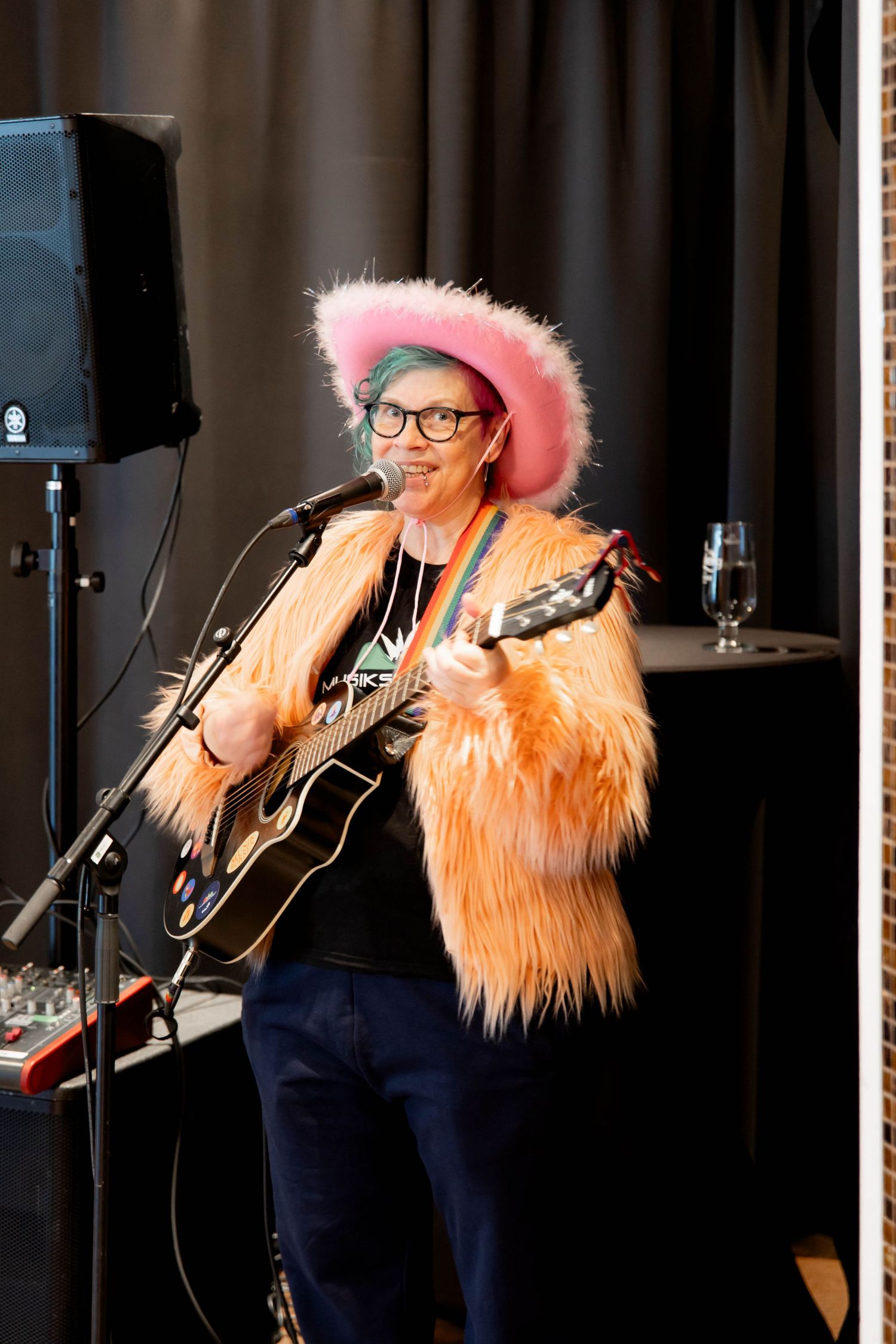 Musician performs on stage wearing a pink fuzzy hat, orange furry jacket and glasses while playing acoustic guitar at microphone.
