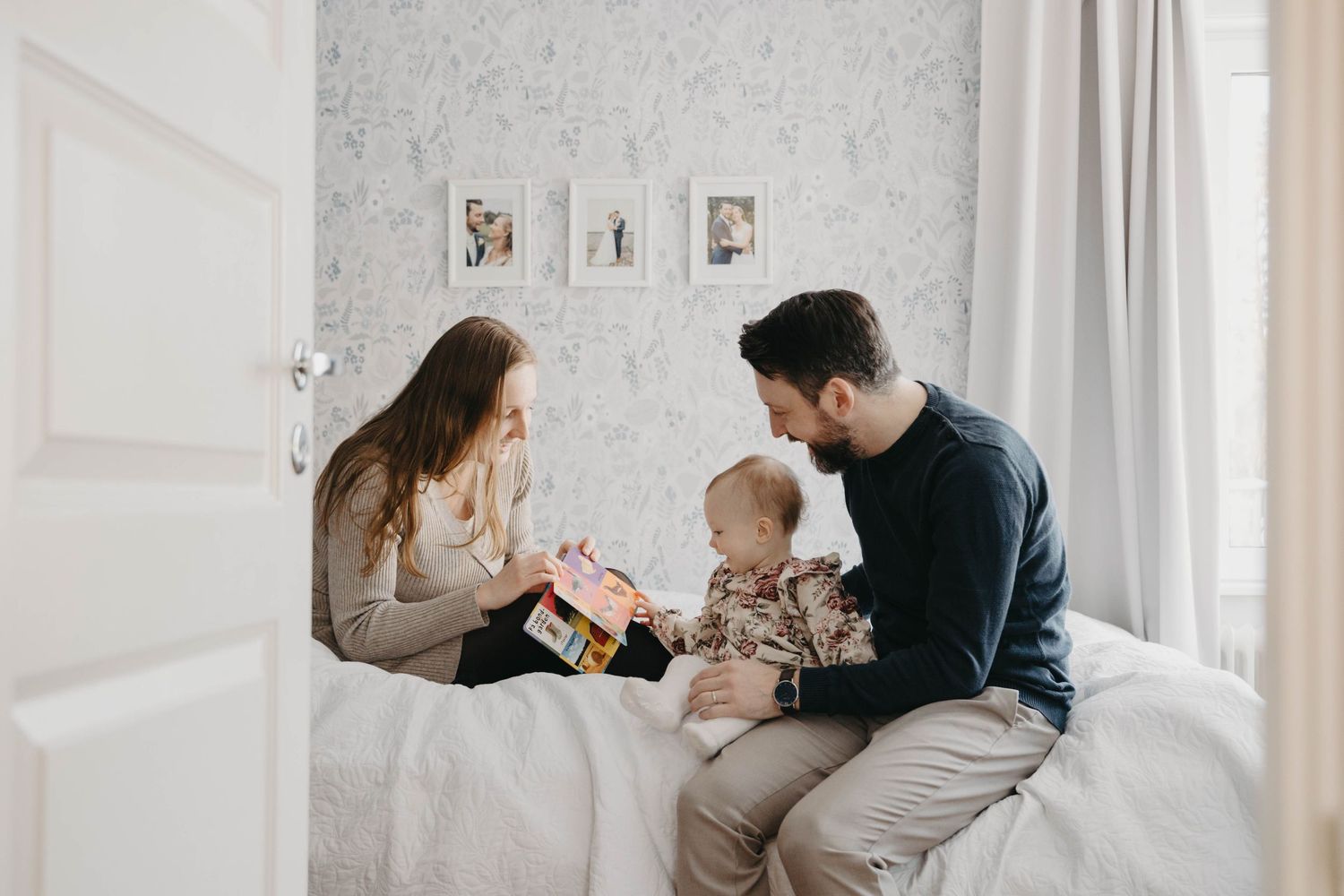 Parents sit on white bed with baby while playing and sharing a tender family moment together.