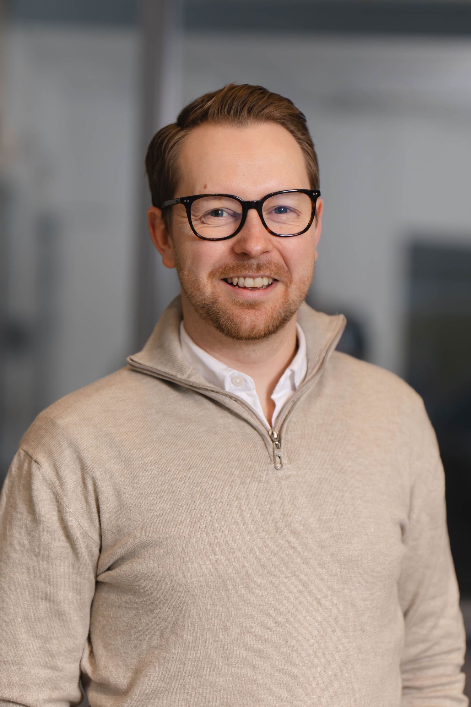 Professional headshot of someone wearing glasses and a beige sweater over white shirt, smiling warmly against blurred background.