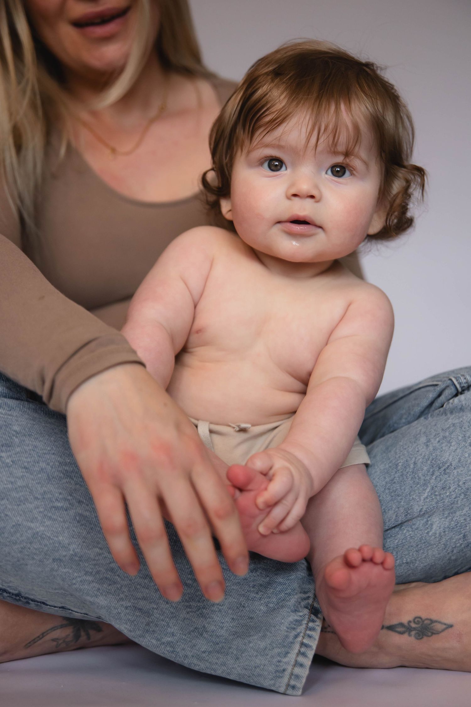 A chubby baby with curly hair sits on a woman's lap, looking curiously at the camera.