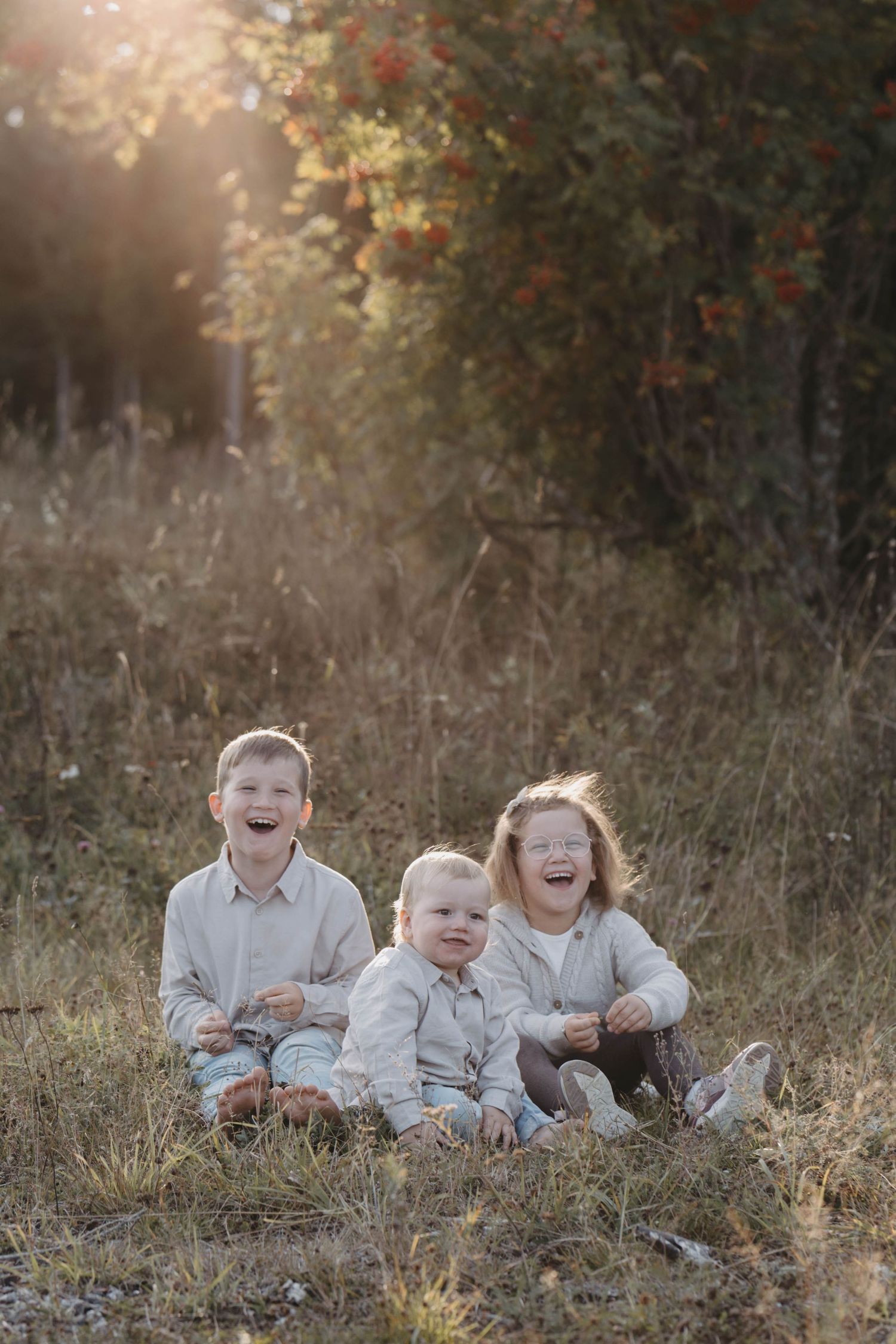 Three children in white clothing sit together in a field during golden hour, sharing joyful laughter and expressions.