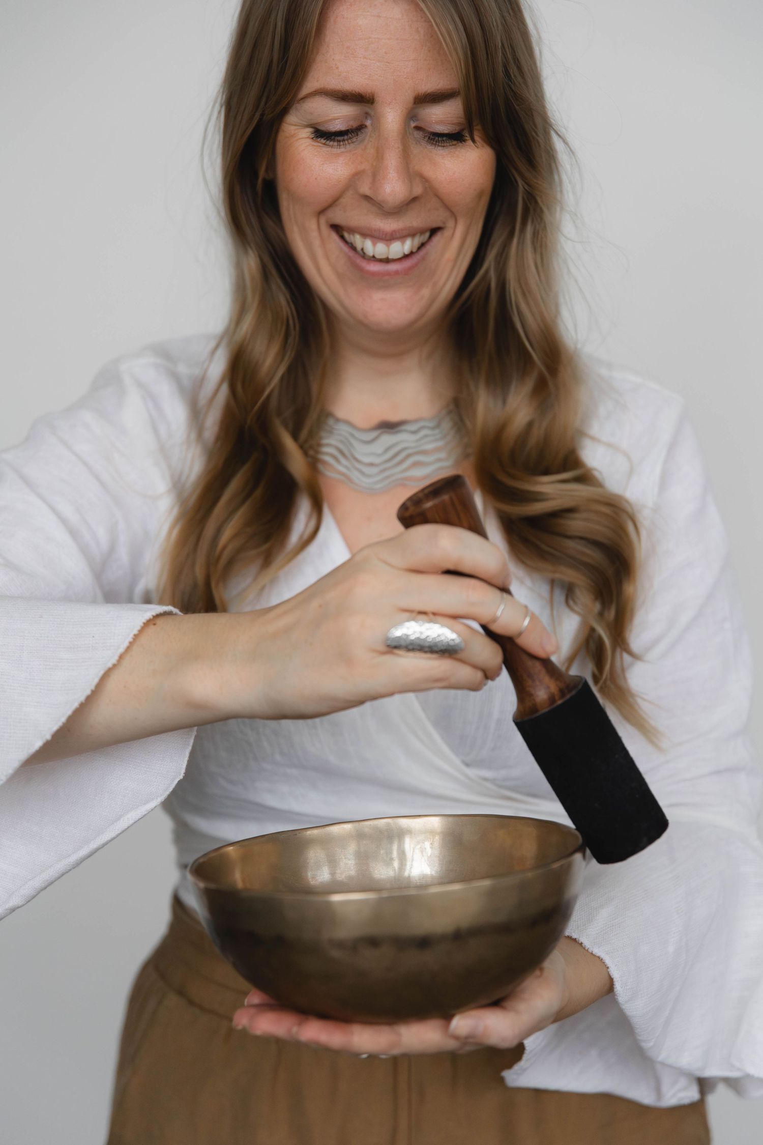 A woman in white holds a meditation bowl and mallet while smiling with eyes closed.