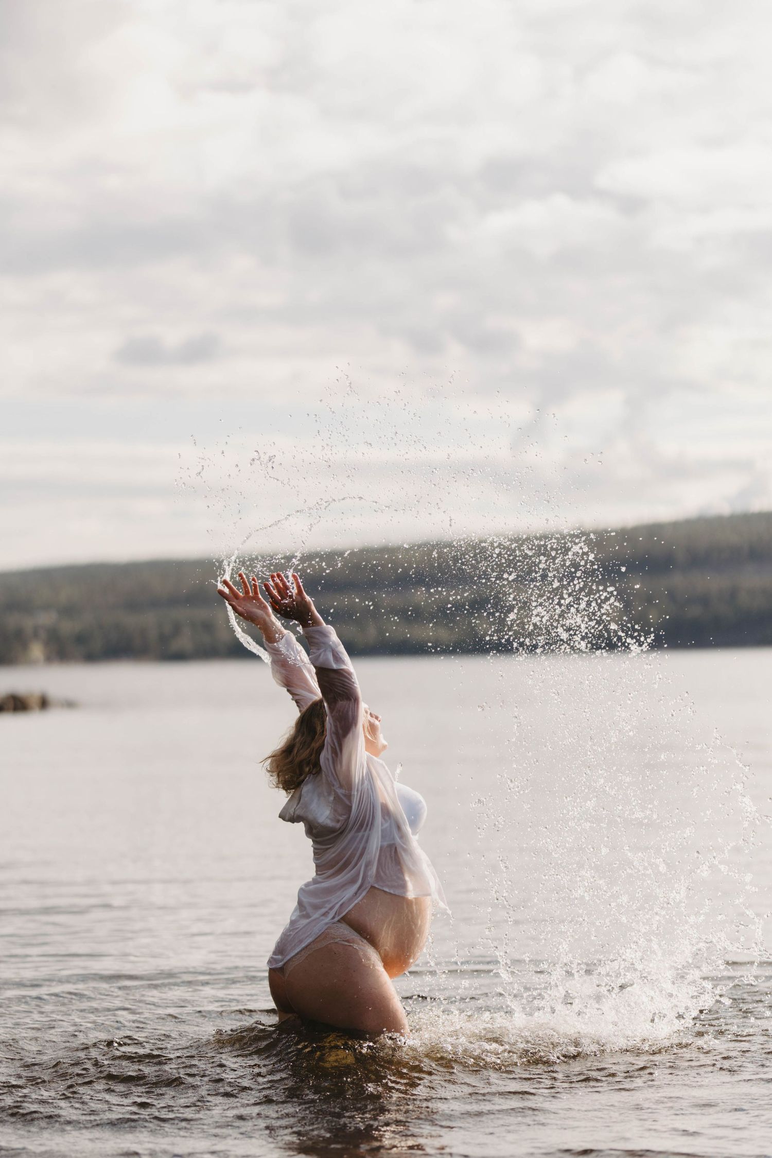 A woman in a white dress splashes and plays in a serene lake during sunset creating joyful water movements.