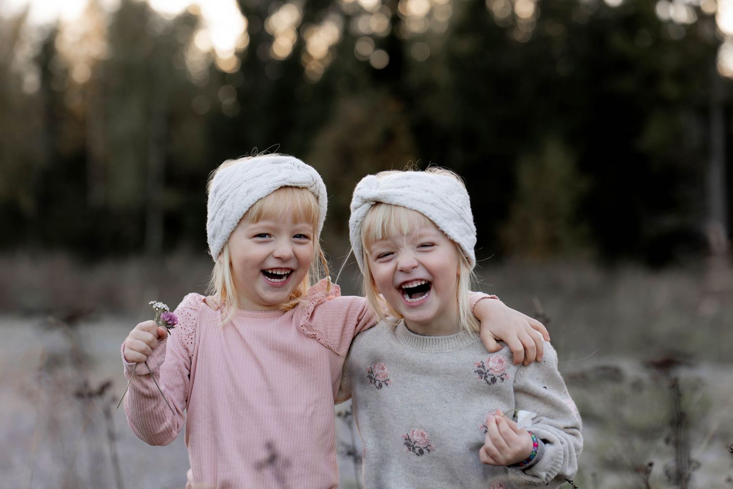 Two children wearing winter hats laugh joyfully together while embracing outdoors in a natural setting.
