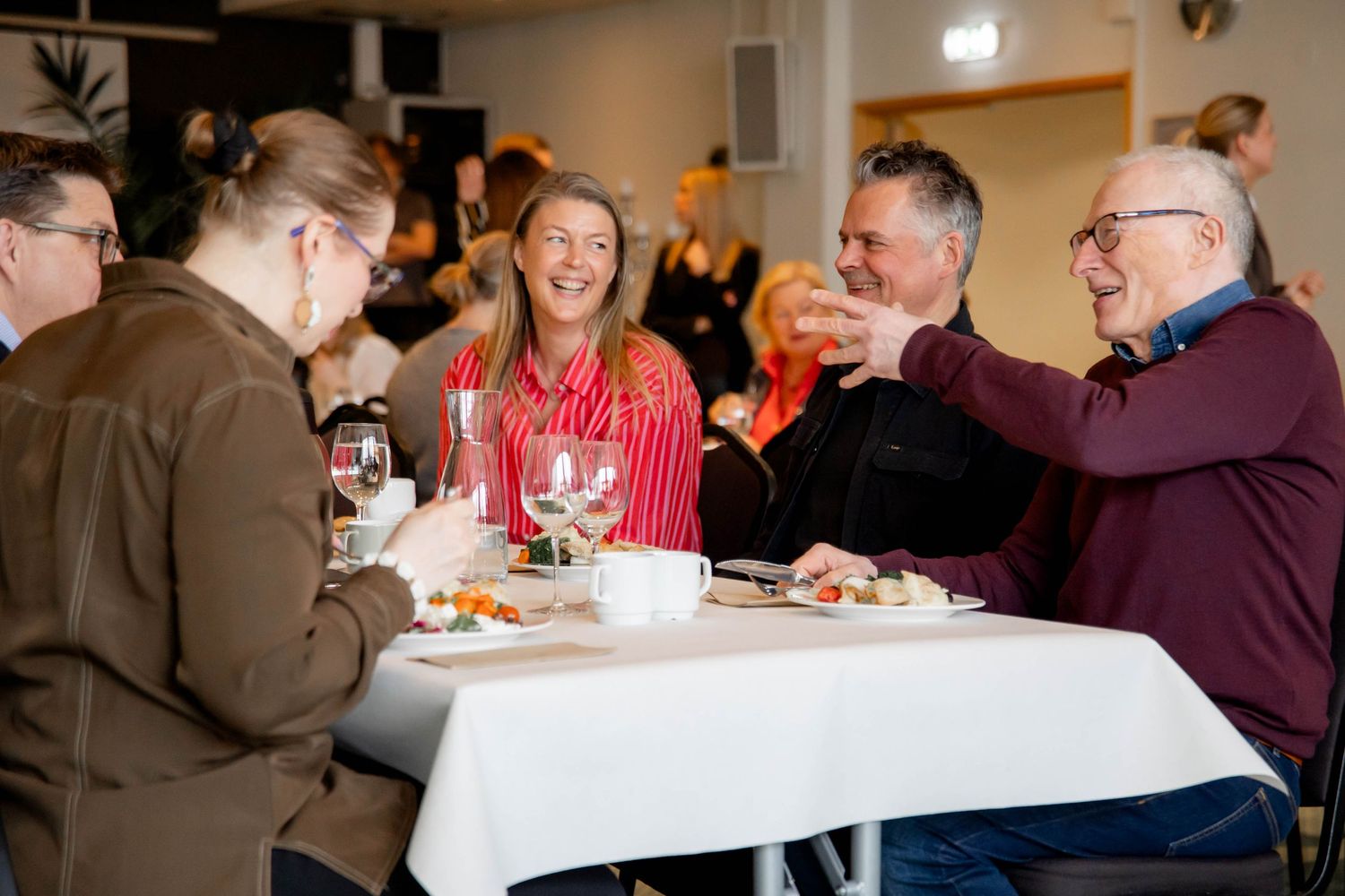 People enjoying a meal and conversation together at a long dining table in a bright indoor setting.