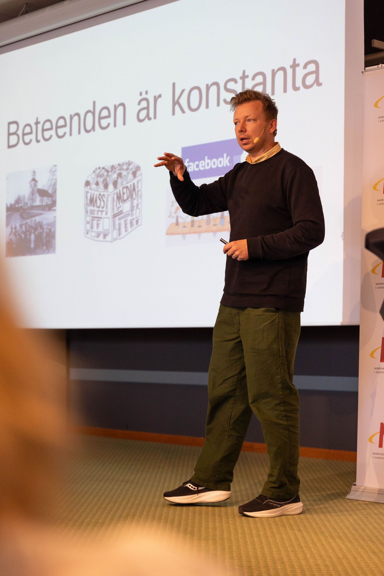 A presenter in a black sweater gestures while giving a presentation with projected slides visible in the background.