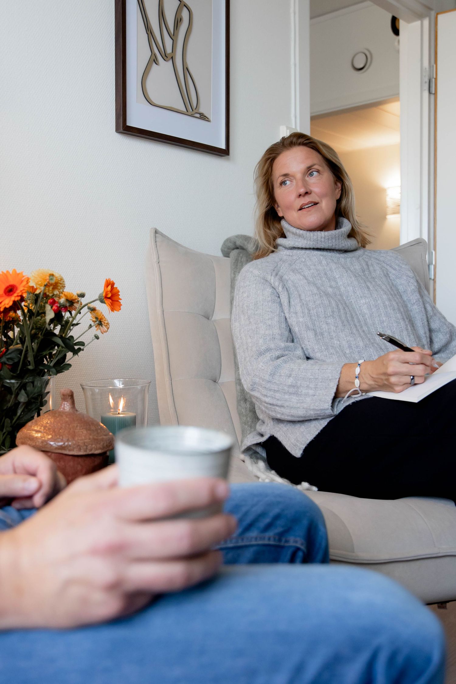 Person in gray sweater relaxing on couch with hot beverage while looking up thoughtfully.