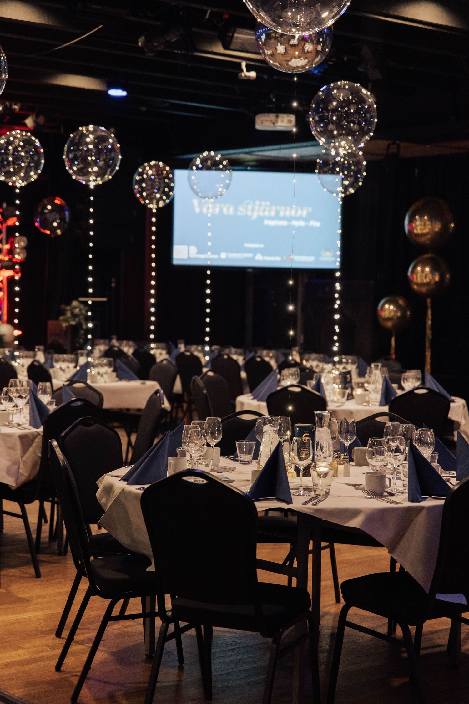 Elegant banquet hall decorated with glowing balloon lights and round tables set for formal dining under a projection screen.
