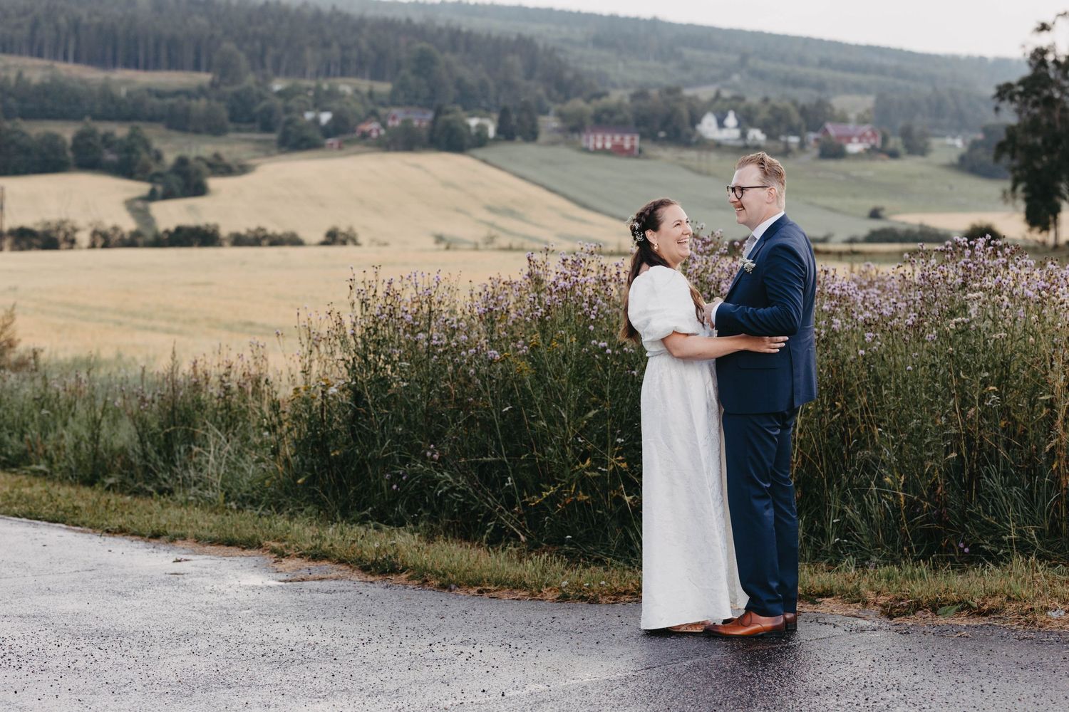 A couple stands together in a scenic countryside field with rolling hills and farmland in the background.