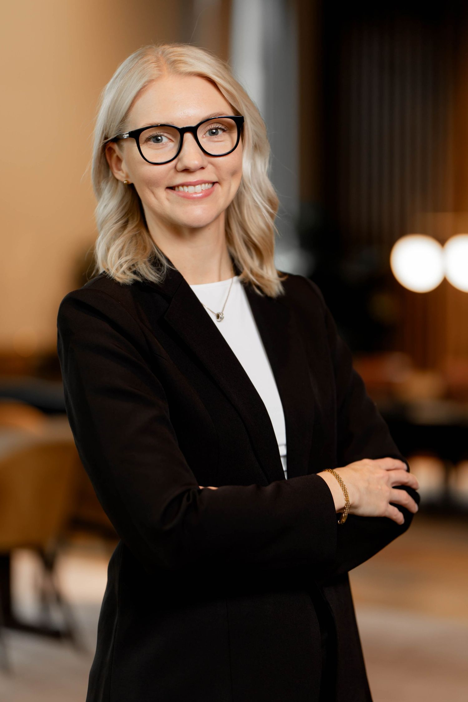 Professional portrait of someone wearing glasses and a black blazer in an office setting with soft lighting in background.