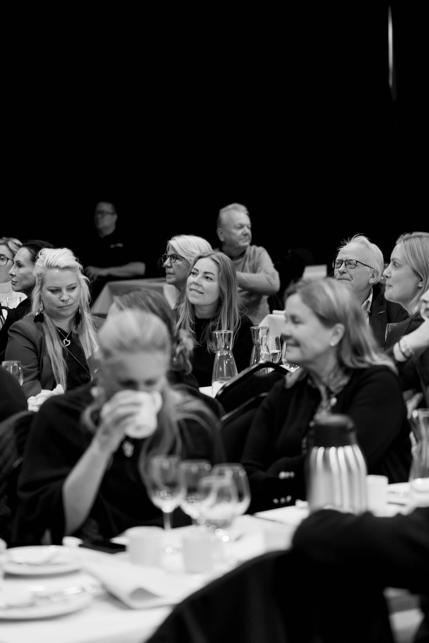 People seated at dining tables engage in conversation during a formal gathering in a dimly lit room.