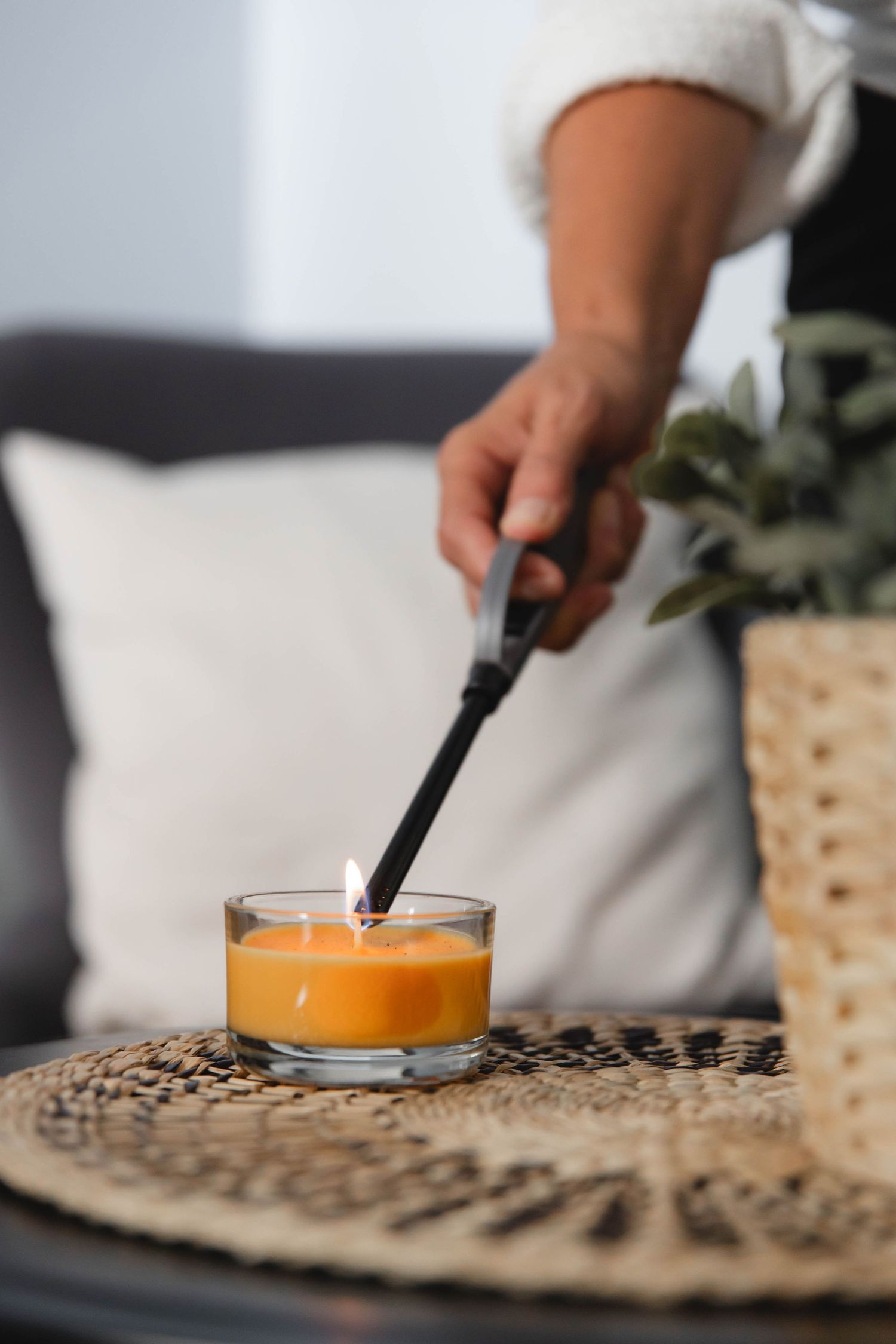 Hand lighting an orange scented candle on a woven placemat with decorative plant in background.