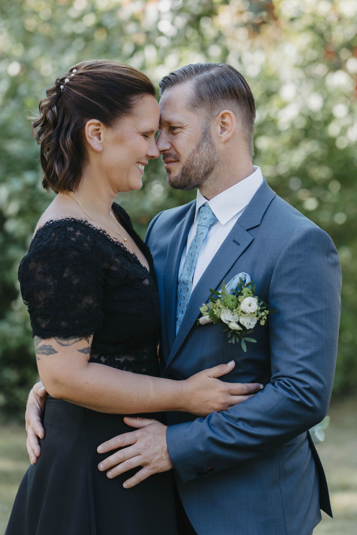 Intimate romantic couple embraces outdoors in formal attire with natural greenery backdrop.