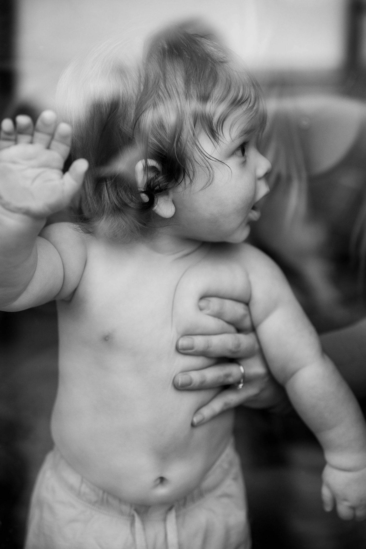 Black and white photo of a shirtless toddler with a hearing aid, held by an adult hand, raising one hand up.